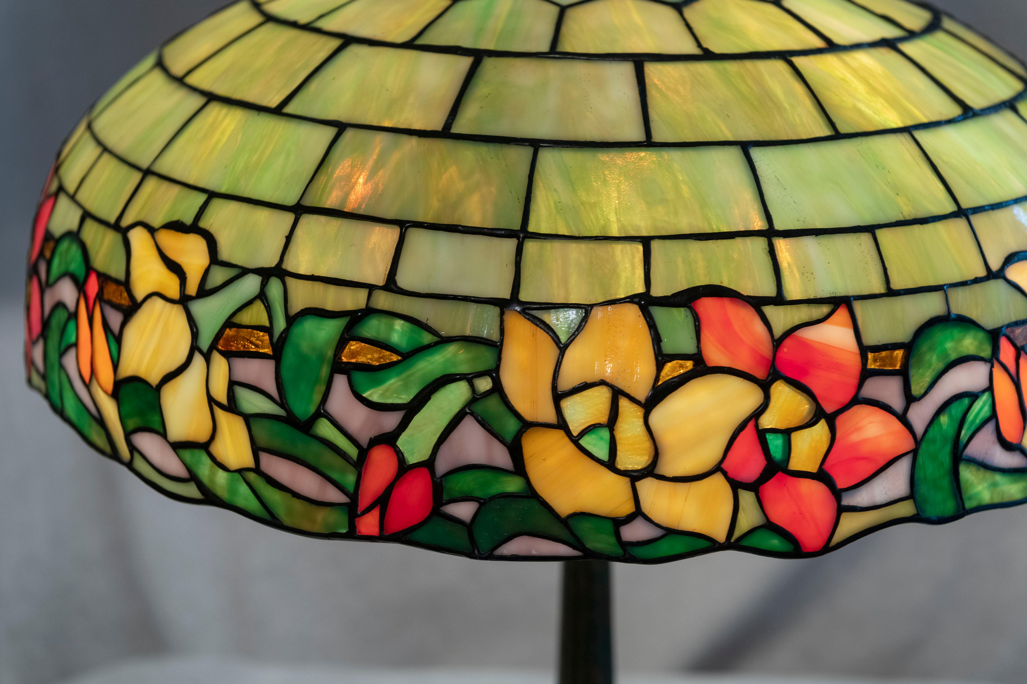 This colorful lamp made by Wilkinson as a graceful floral border of orange, green, yellow and purple tiles. The upper geometric brickwork design section is comprised of yellowish-green tiles. The handsome base is factory patinated in a rich dark