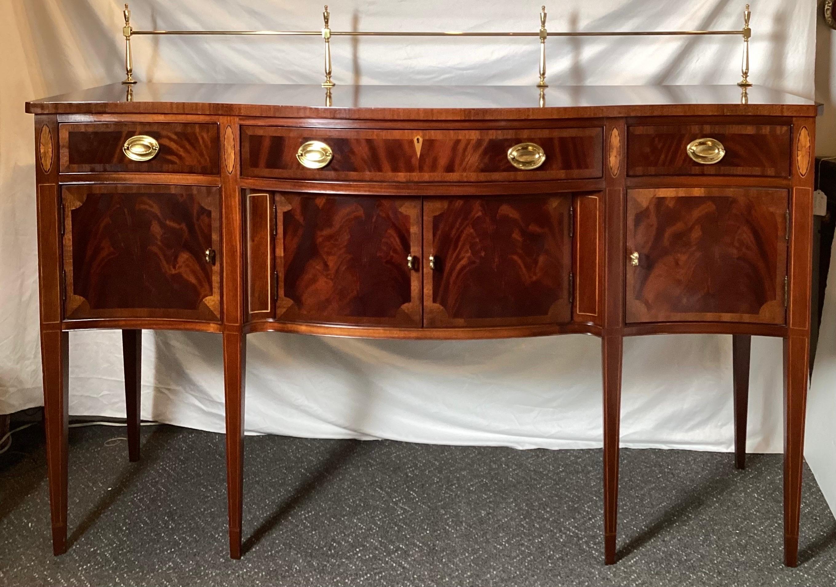 A classic Hepplewhite style mahogany inlaid side board. The shapely front with cross banding with inlaid medallions on tapering pencil inlaid legs. The top with an 8.5 inch tall removable brass brandy gallery railing.