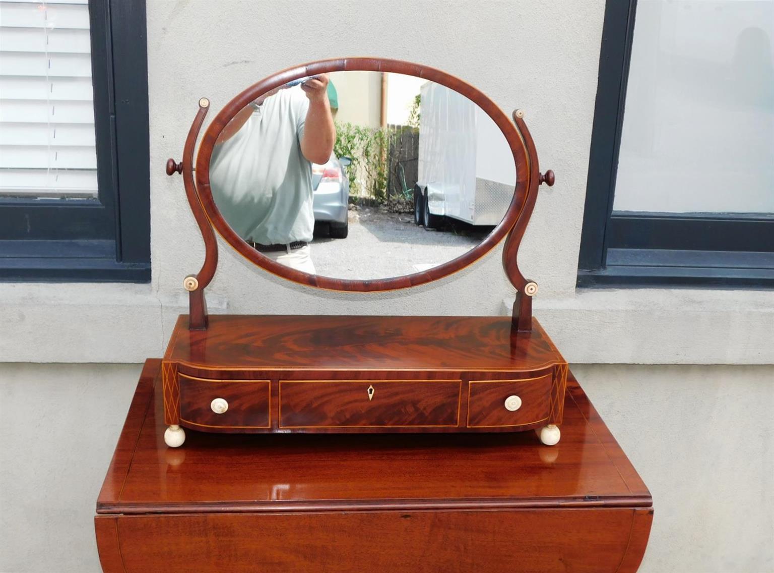 American mahogany bow front three drawer oval dressing mirror with flanking serpentine mahogany supports, circular reeded bone medallions, satinwood string inlays, crossbanded border, flanking bone drawer pulls, central diamond bone inlaid