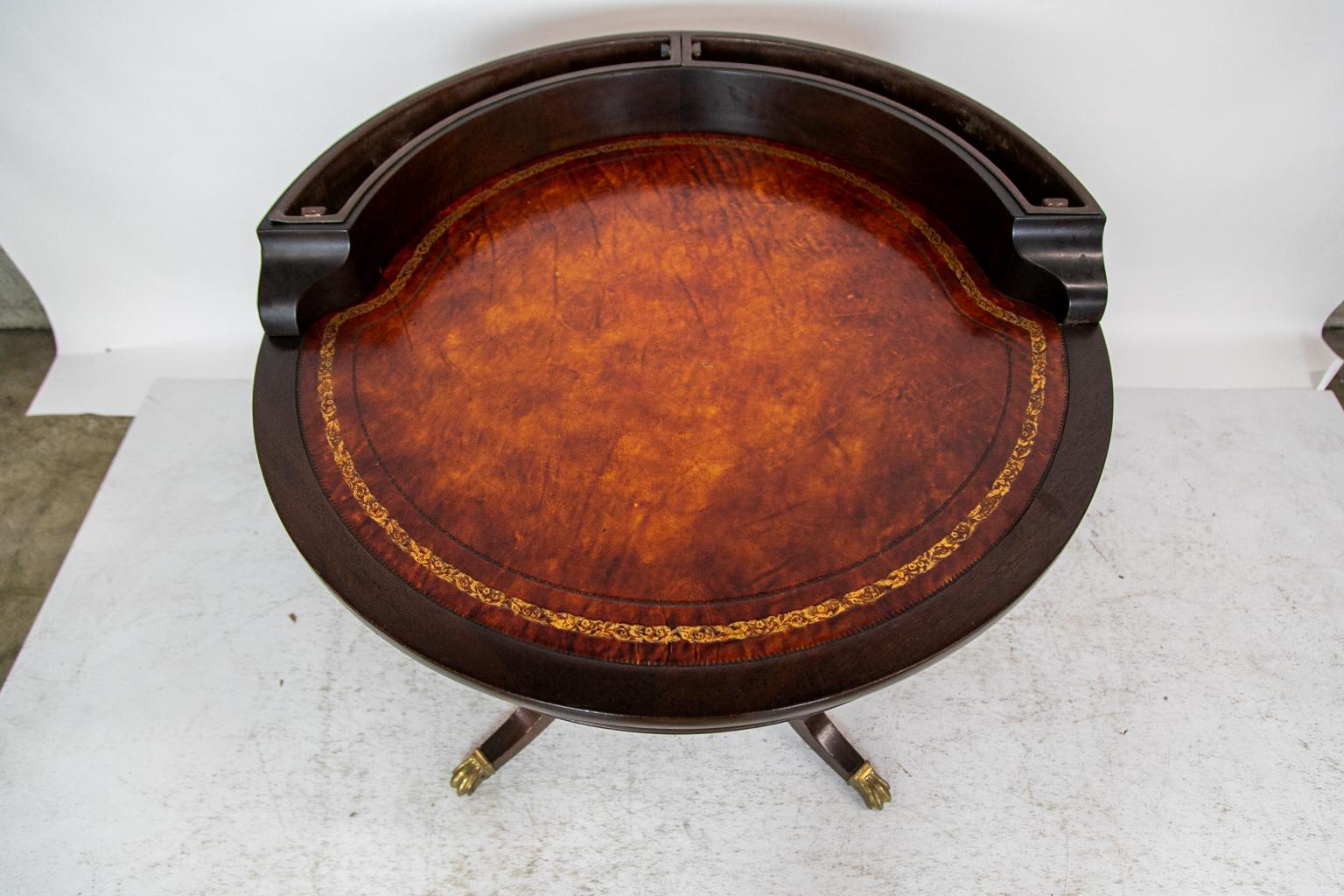 This drum table has a burnt orange leather top with a large gold tooled border flanked by a blind tooled inner border and outer edge. The gallery has two removable liners. The platform base has the original cast brass medallions.