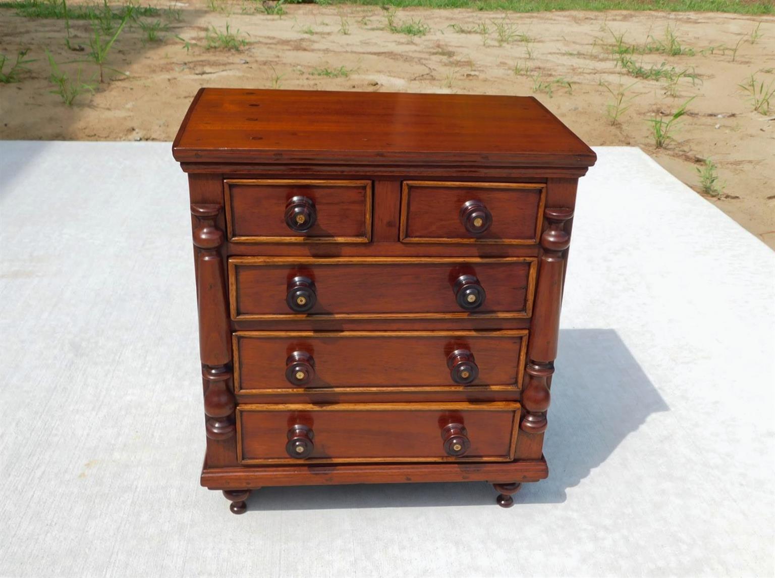 American Sheraton Mahogany five drawer miniature chest with a carved molded edge top, flanking turned bulbous half columns, turned wooden knobs with central mother of pearl inlays, and resting on a molded edge skirt with the original finial bun