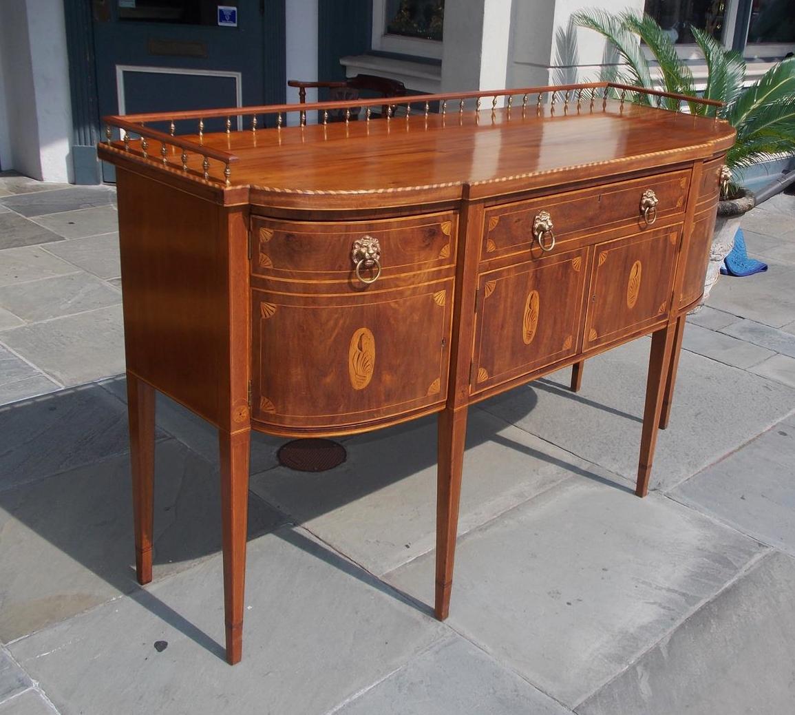 American Hepplewhite style mahogany sideboard with an acorn brass and mahogany rail gallery supported by a curvature and straight inlaid holly top, centered patera inlaid drawer over two lower conch shell inlaid cabinet drawers, original lions head