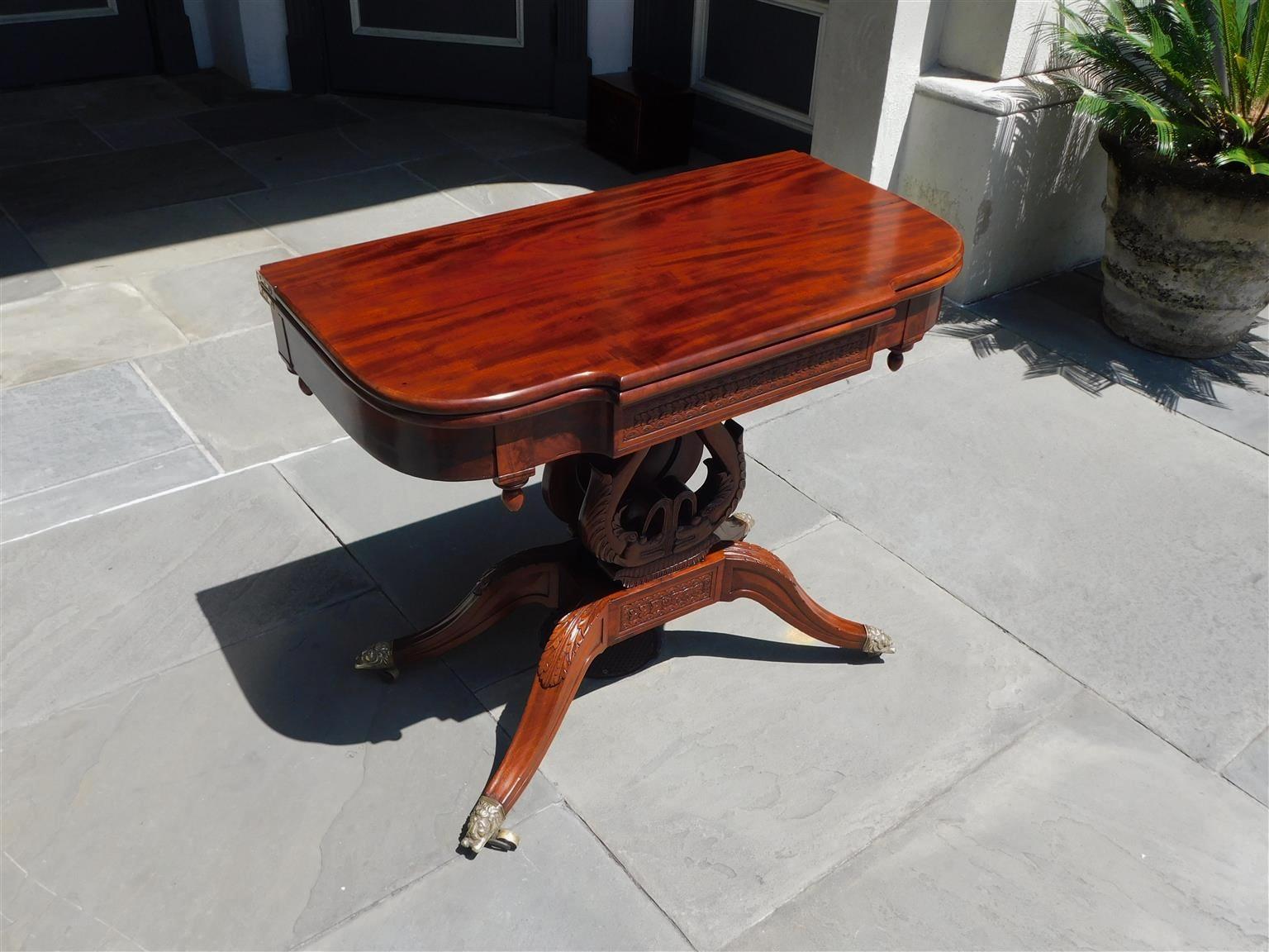 American neoclassical cuban mahogany elliptical game table with a swivel hinged curvature top, decorative geometrical carved figured apron, flanking carved corner acorns, lyre form base with intertwined dolphins spouting water, and resting on