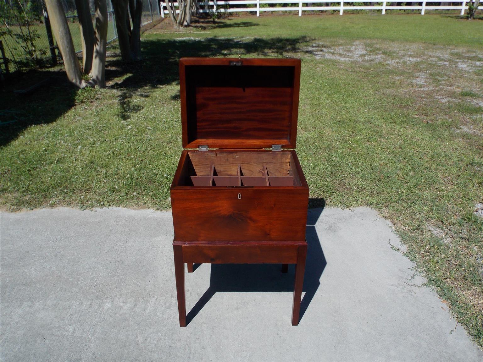 American mahogany hinged bottle box on stand with exposed dovetails, interior dividers, original brass side handles, and centered brass knob with original locking key. Early 19th century.