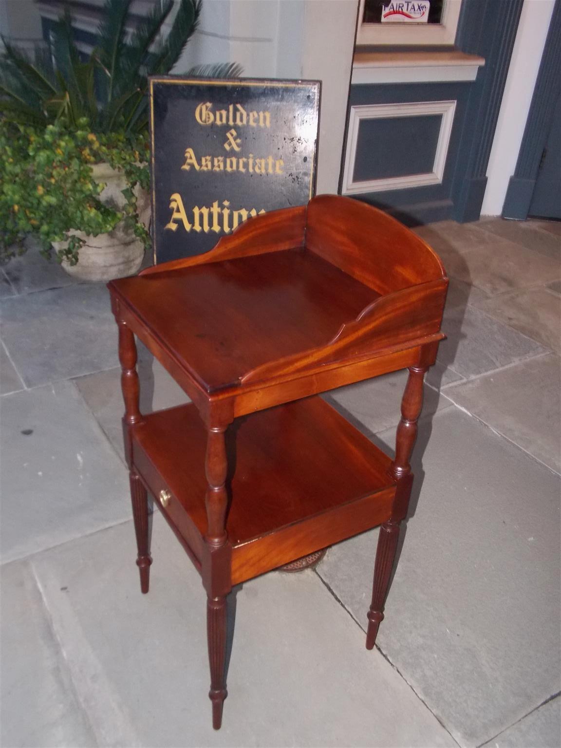 American mahogany Sheraton wash stand with a upper back splash, centered interior holes for water basin and shaving cups, turned bulbous ringed columns, lower shelf over one drawer with brass pull, and terminating on the original reeded ringed legs.
