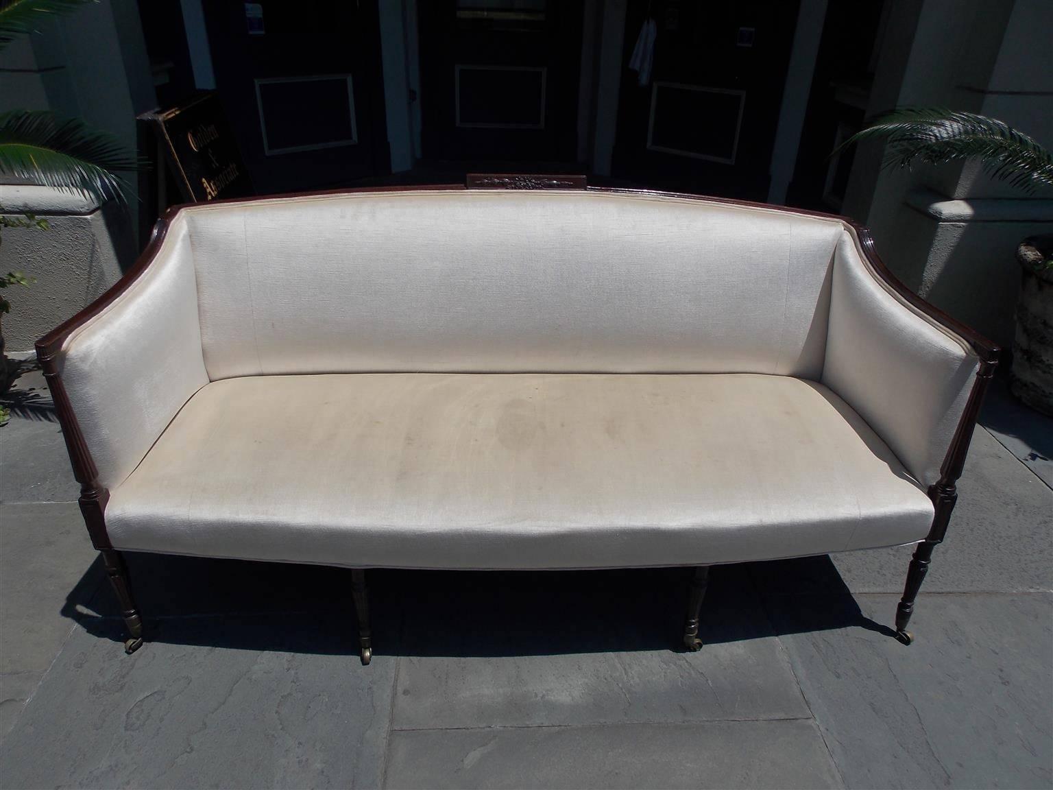 American mahogany Sheraton style sofa with an arched back , centered carved ribbon with graduated bell flower cartouche , sweeping scrolled reeded arms, and terminating on reeded squared tapered legs with the original brass casters. Late 19th