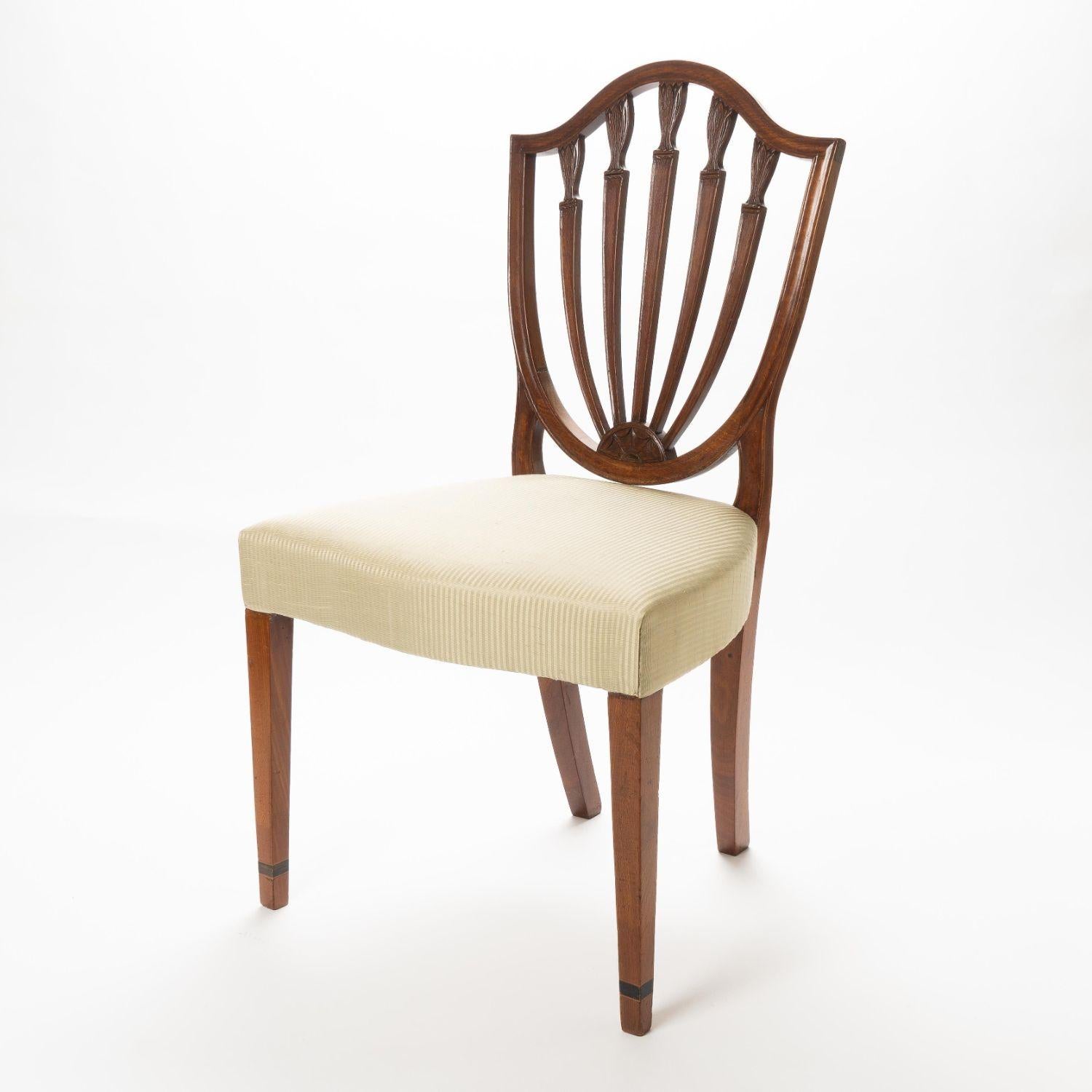 American mahogany shield back side chair with over rail upholstered serpentine seat. The four vertical back rails are carved with water leaf at the point they join the serpentine crest rail. The base of the heart back is a carved demi-lune. The