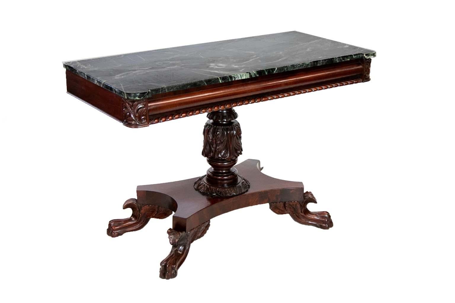 The front of this mahogany American empire console table has a cushion frieze above gadrooned molding. The corners have carved stylized acanthus leaves. The supporting stem has acanthus leaves carved in high relief. A platform base has concave edges