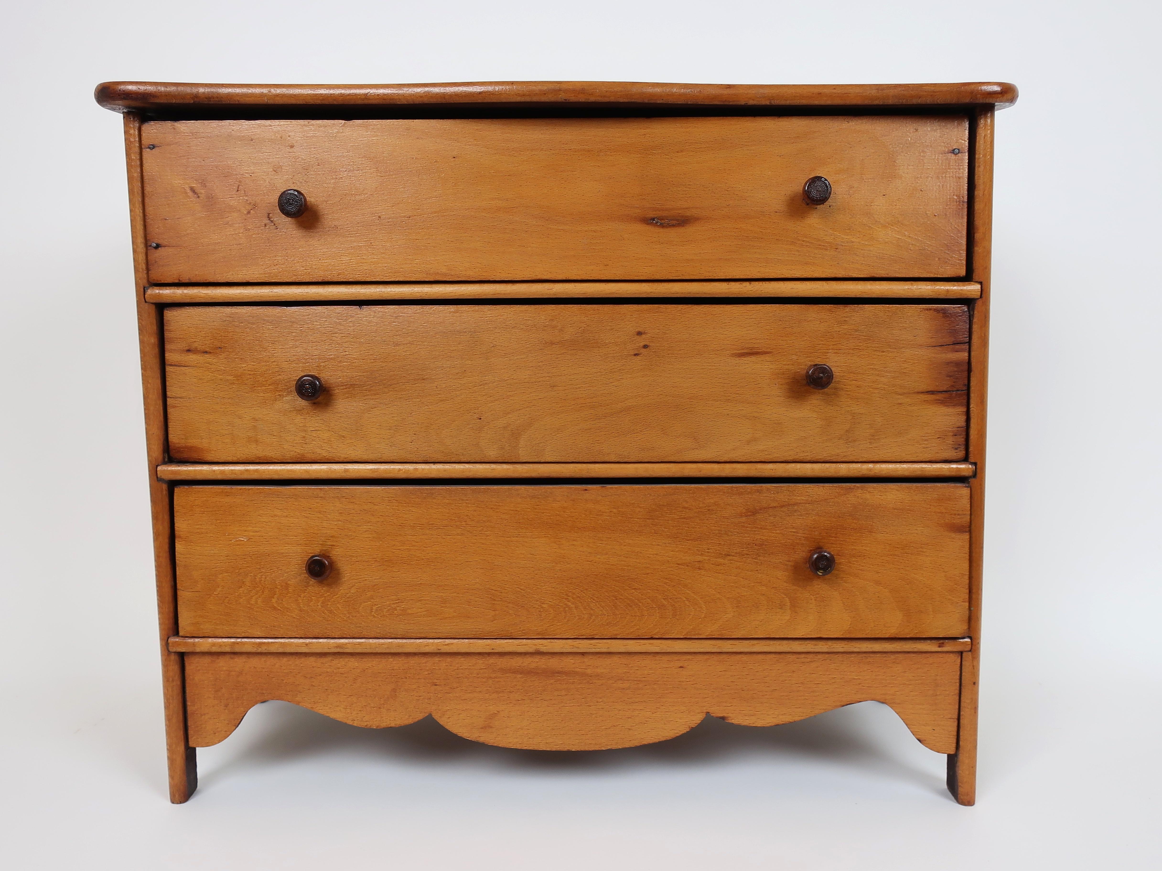 The charming miniature chest is pine. It has three drawers that are of dovetail construction and the chest has turned wood knobs. The top is shaped and the chest has a scalloped apron on the front and sides. It is in very good condition.