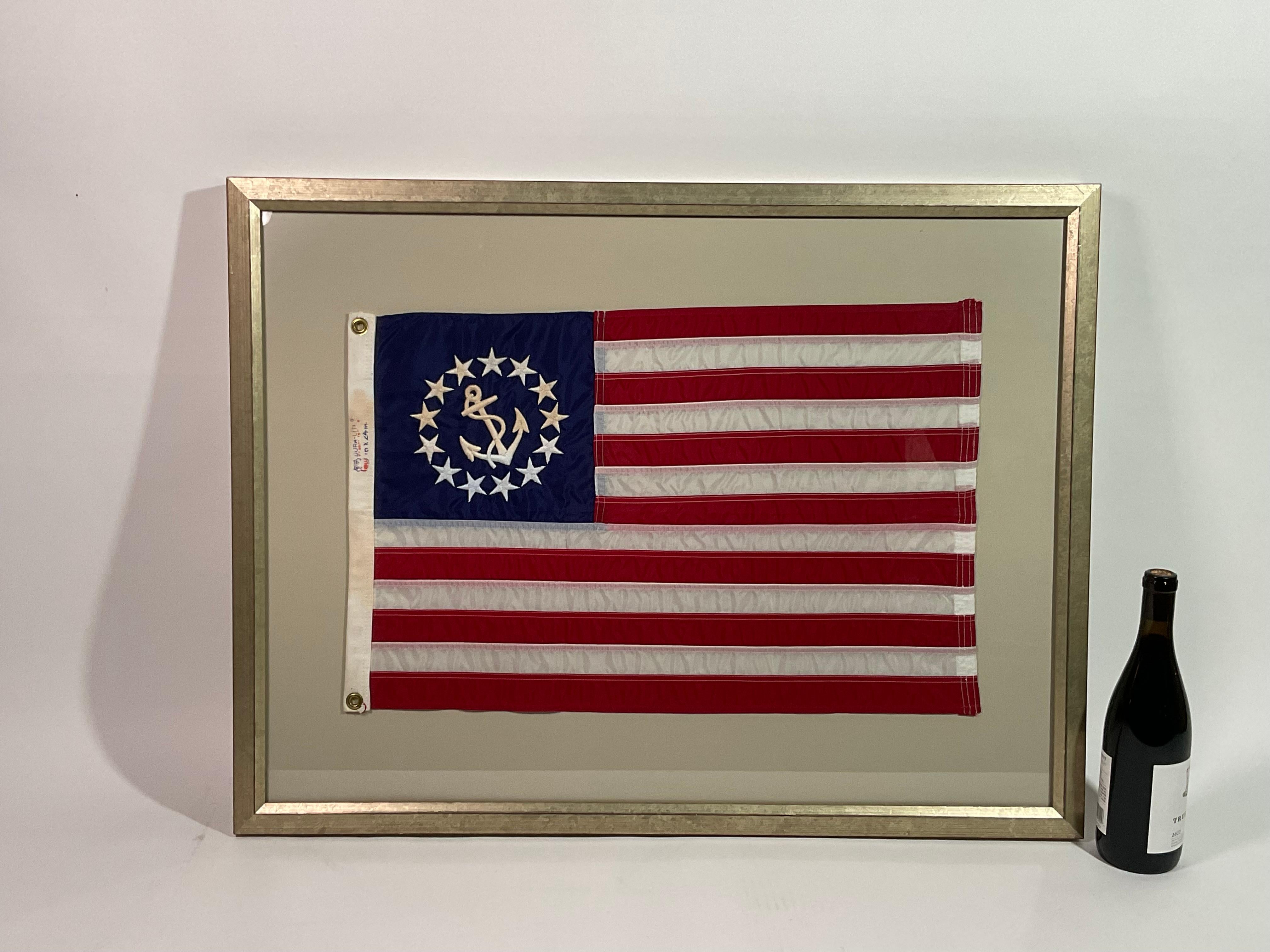 Bandera de enseña de yate estadounidense enmarcada. Bandera náutica con un panel azul con un ancla fouled bordada y trece estrellas, y franjas rojas y blancas con un izado de lona y ojales de latón. Montado sobre paspartú beige y ajustado a un marco