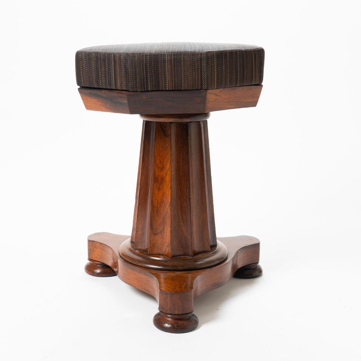 American neoclassic upholstered seat piano stool. The seat rests on a fluted and truncated columnar pedestal with trefoil platform base on turned and suppressed bun feet. Rosewood and rosewood veneers on eastern white pine. The seat is newly