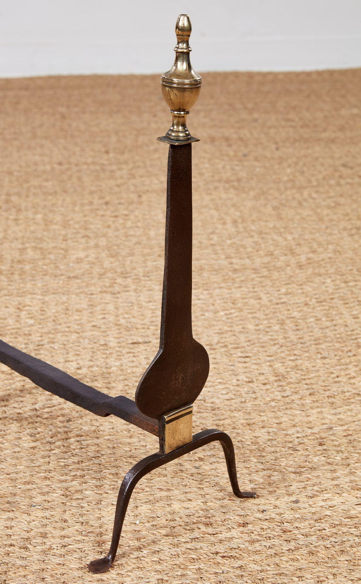 A pair of neoclassical andirons having blacksmith forged flat 'knife blade' shafts with shaped silhouettes, on fine arched feet below a decorative brass plate, and topped with turned brass finials.  American, circa 1810.
