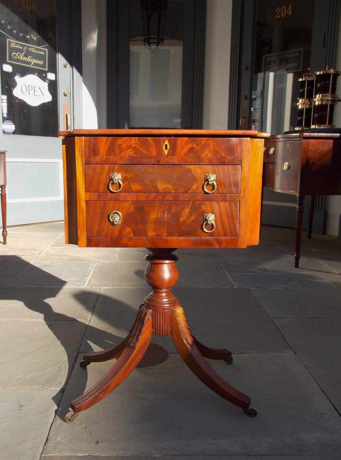American neoclassical mahogany hinged two-drawer work table with canted corners, original lion head brasses, fitted interior compartments, diamond escutcheon, turned bulbous ringed pedestal, and terminating on acanthus carved reeded saber legs with