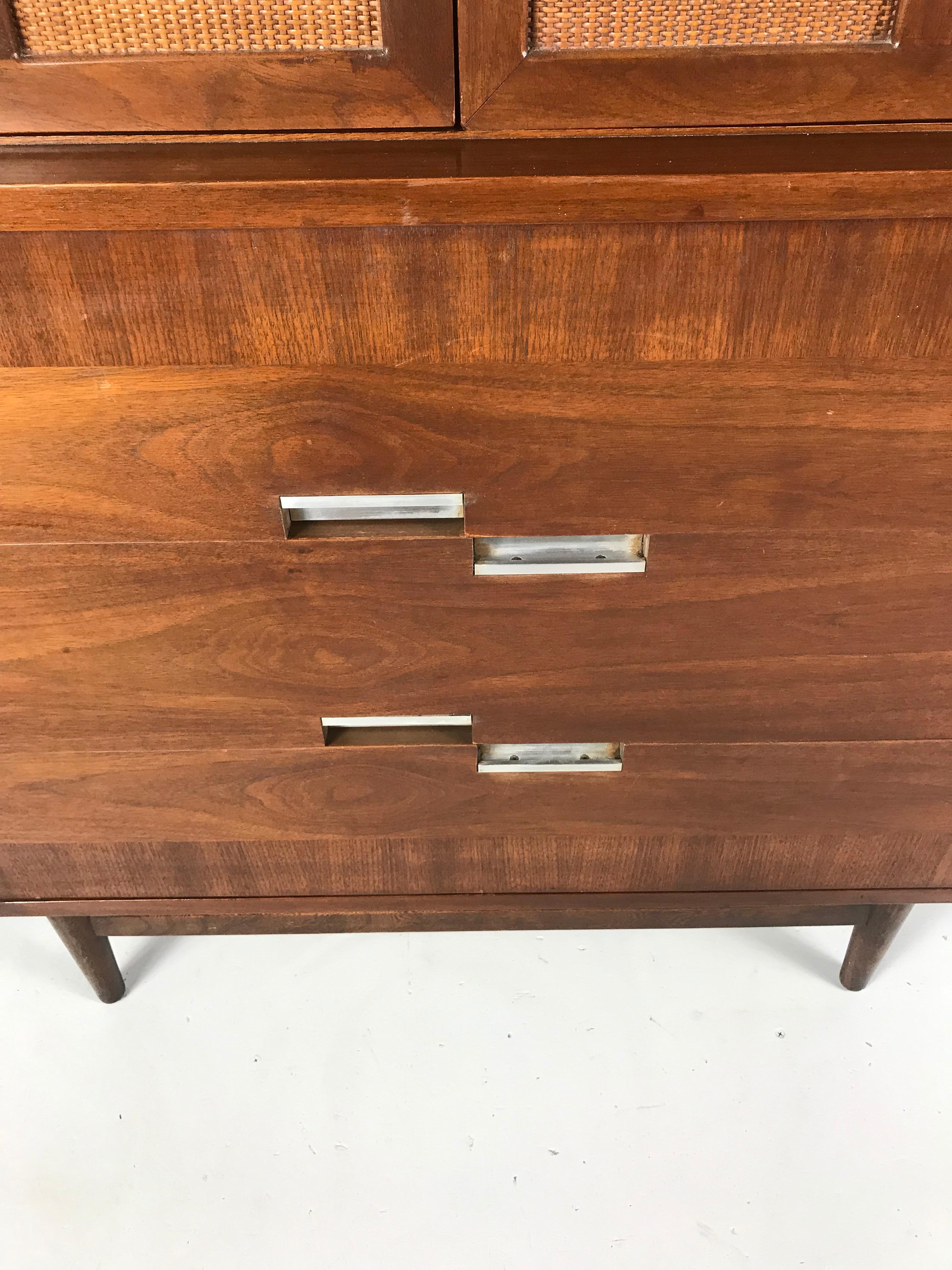 This stylized dresser features contrasting walnut veneer, two doors with four rattan panels, and this lines signature inset nickel “x” on each corner of the top. Rich walnut grain graces all surfaces. With an angular profile, this vintage piece has