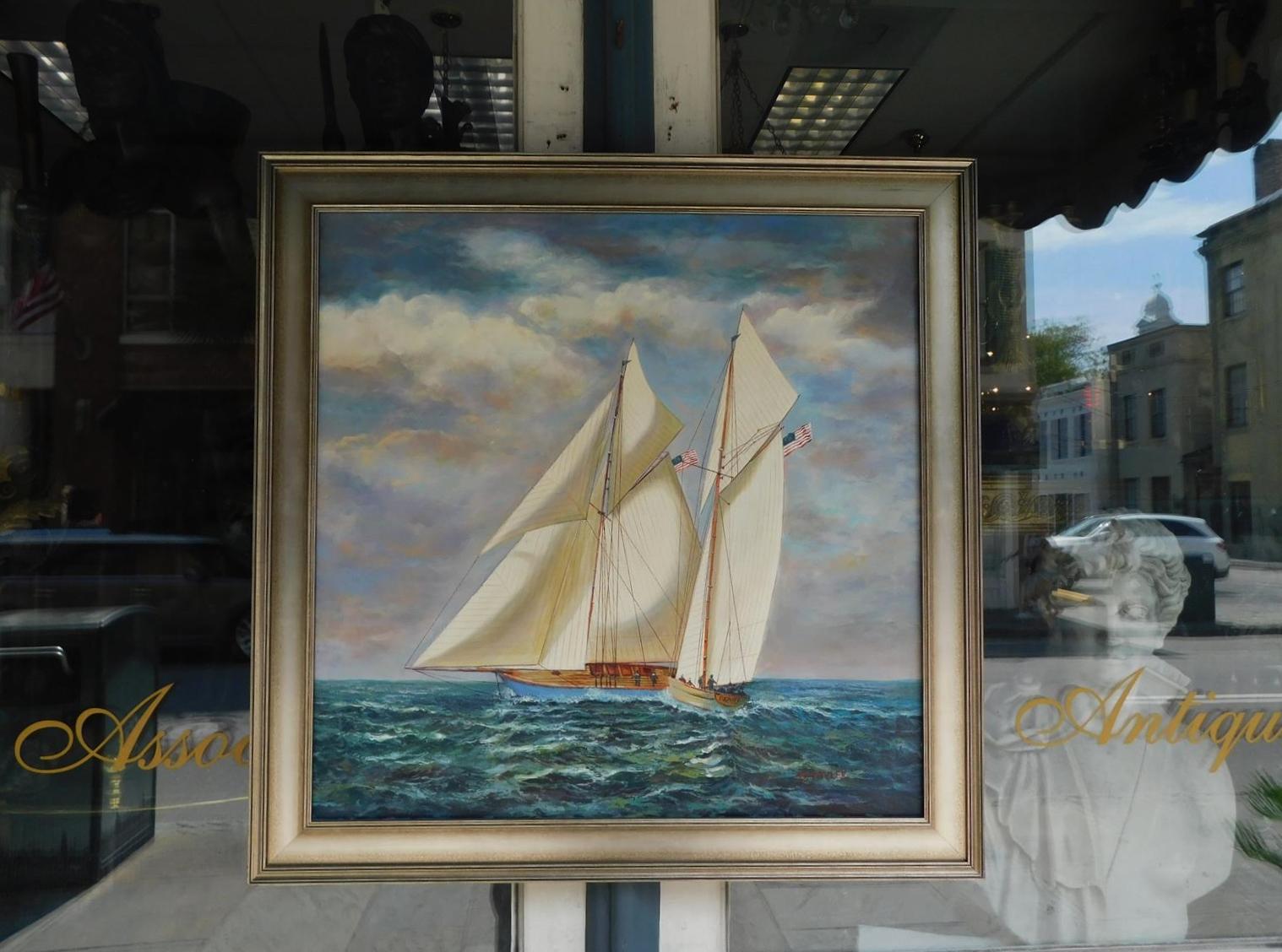 American nautical oil on canvas of single masted schooner yacht under full sail with American flags, and crews on deck while second vessel victory is approaching.  Painting is mounted in the original silver gilt frame, Early 20th century. Signed D.