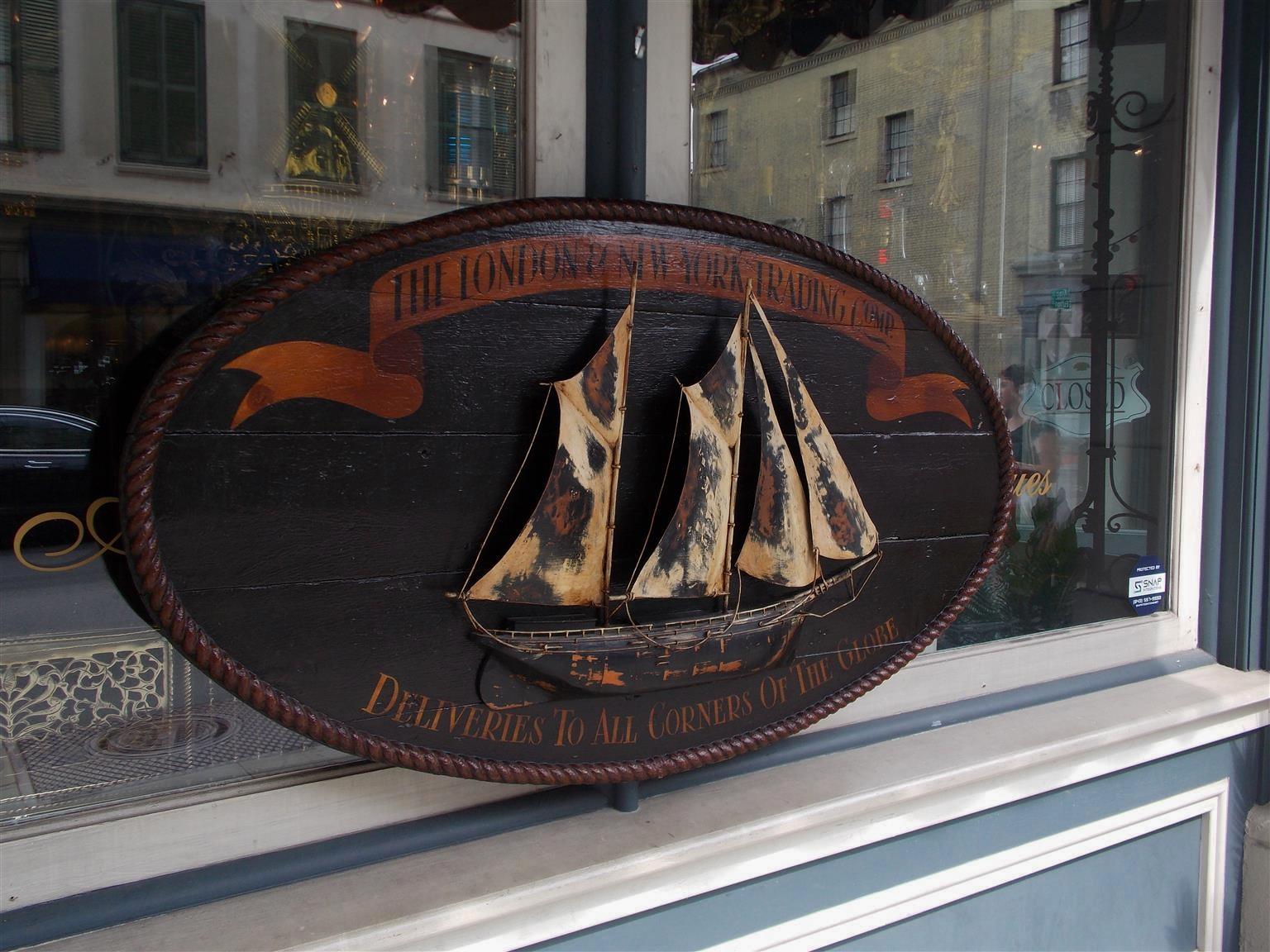 American oval hand carved timber trade sign with a centered two masted ship, rope border, upper painted banner 