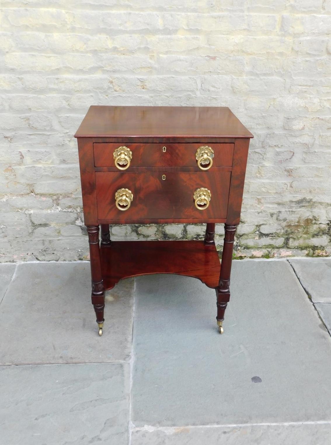 American Regency mahogany side table with a carved molded edge top,  two graduated drawers, original foliage medallion brass pulls, four foliage ringed support columns with an arched lower shelf, and resting on tapered foliage feet with the original