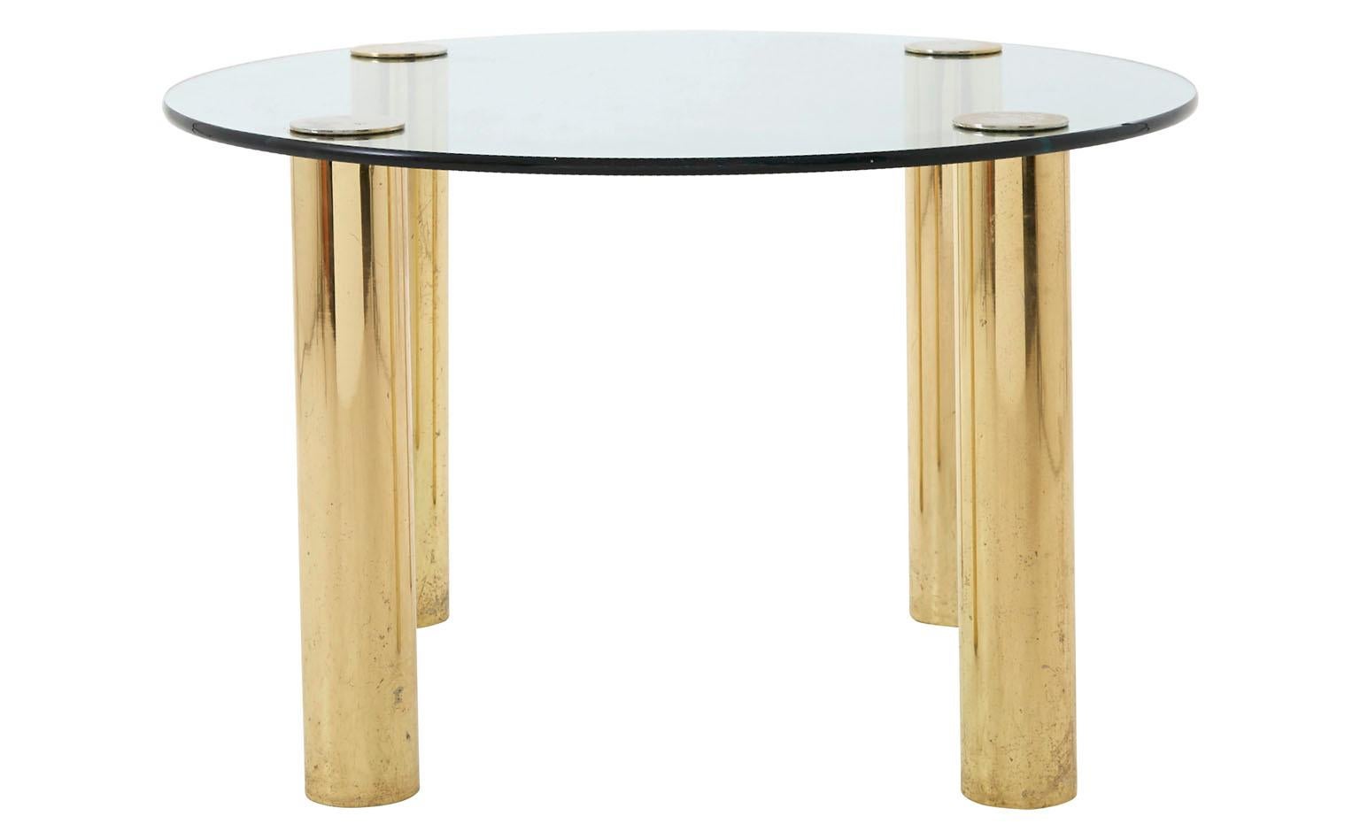 Our Hollywood Regency-style Vintage Brass Dining Table was made in America in the 20th century. It features its original round glass top, supported by four cylindrical, shiny brass legs. It is in good condition, with some scratches and wear