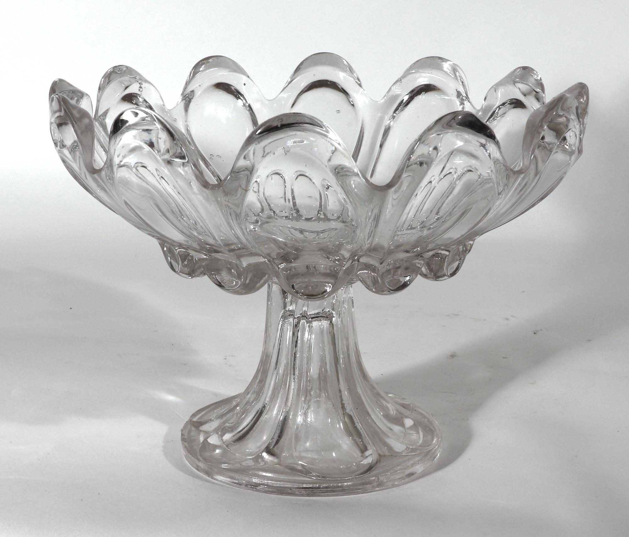 Verre sandwich américain compote lobée
Milieu du XIXe siècle

Le compotier circulaire à pied en verre avec douze lobes formant un bord ondulé. La cuvette supérieure repose sur un pied évasé creux.

Dimensions : 7 pouces de haut x 9 1/4 pouces