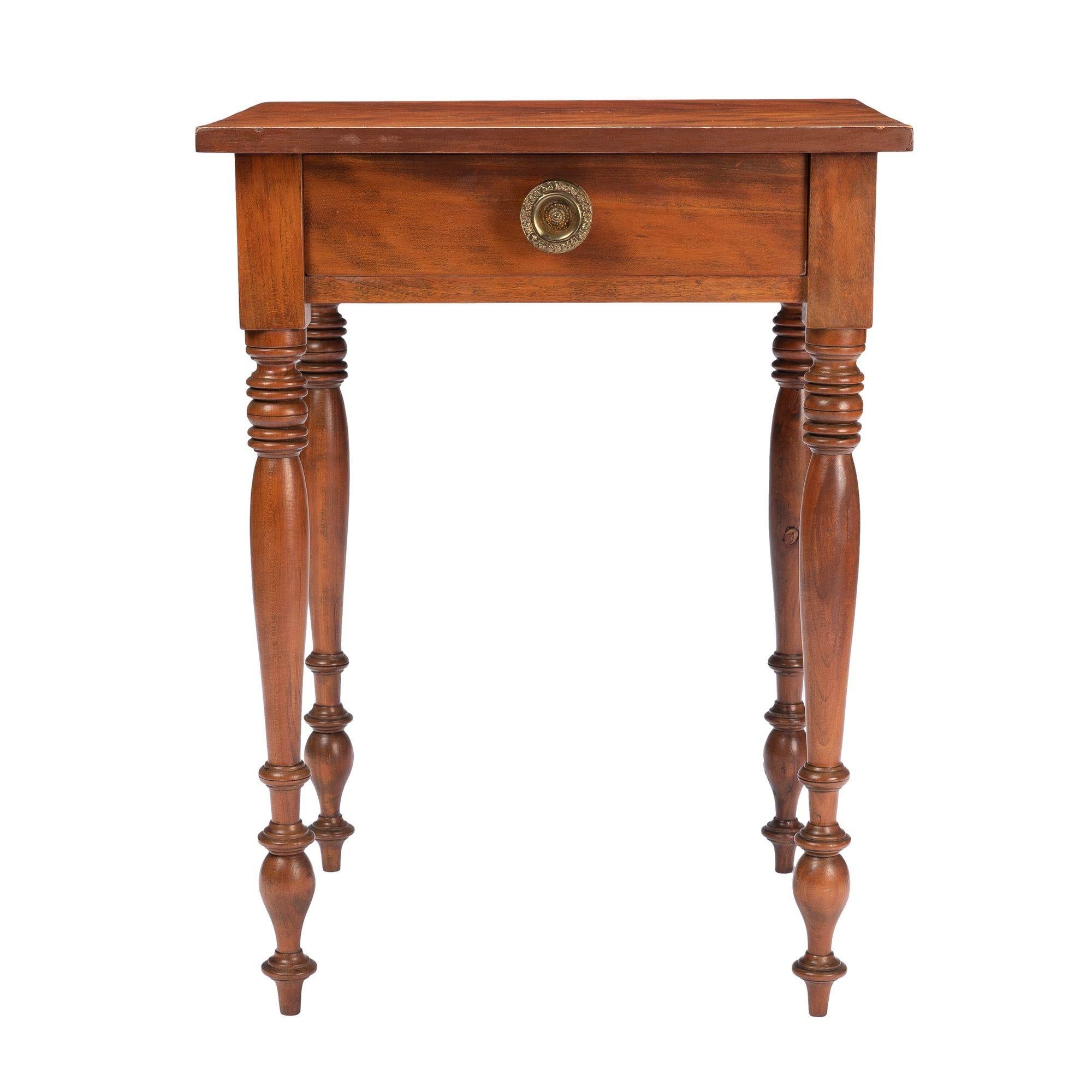 This American Sheraton curly cherry one drawer stand features a single board top on a conforming apron with a single drawer, fitted with its original stamped brass drawer pull. The corner dies continue into vigorously turned legs terminating in a