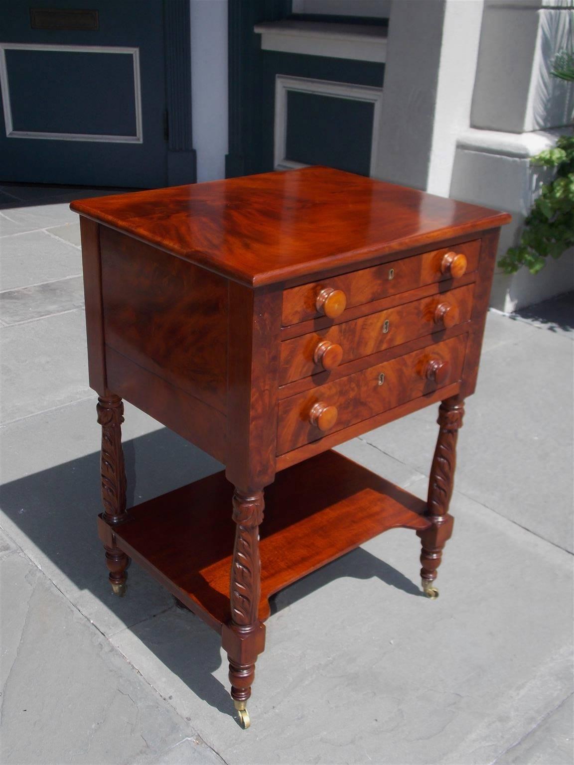 American Sheraton flame mahogany side table with a carved molded edge top, three graduated drawers with centered key hole escutcheons, original wooden knobs, flanking flame mahogany blocked corners, lower connecting shelf, and terminating on