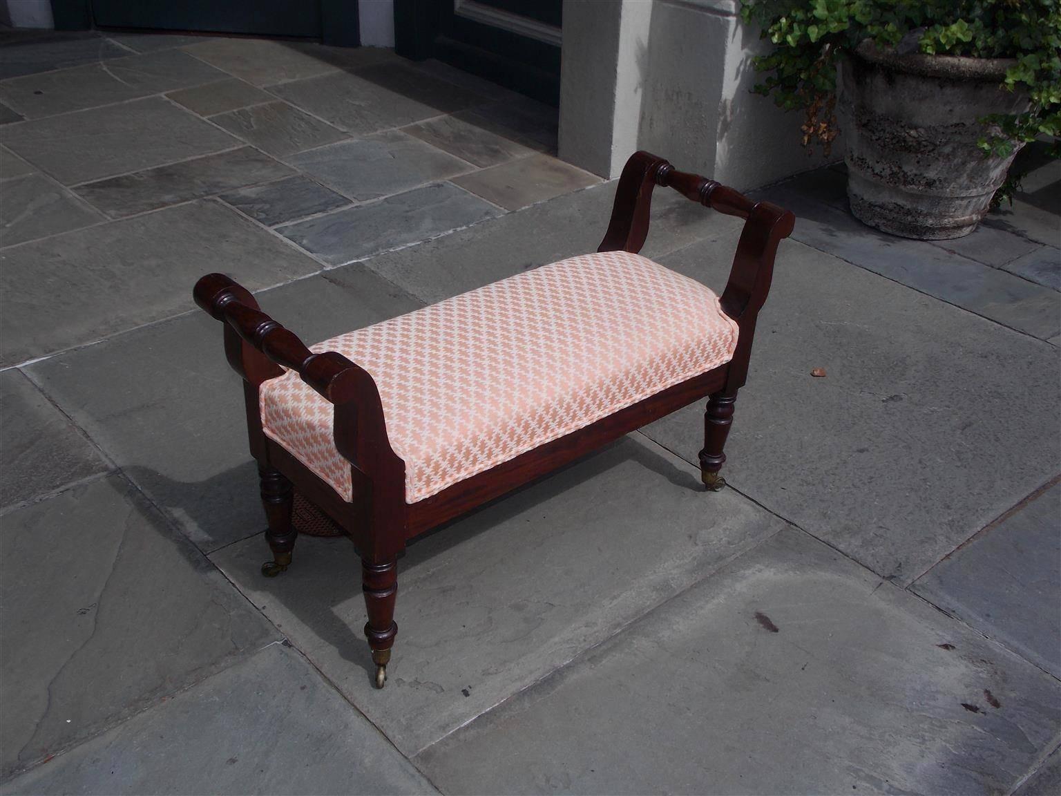 American Sheraton mahogany upholstered window bench with flanking scrolled arms, carved molded skirt, and terminating on turned bulbous ringed legs with the original brass casters. Baltimore, Early 19th century. Bench can be reupholstered at an
