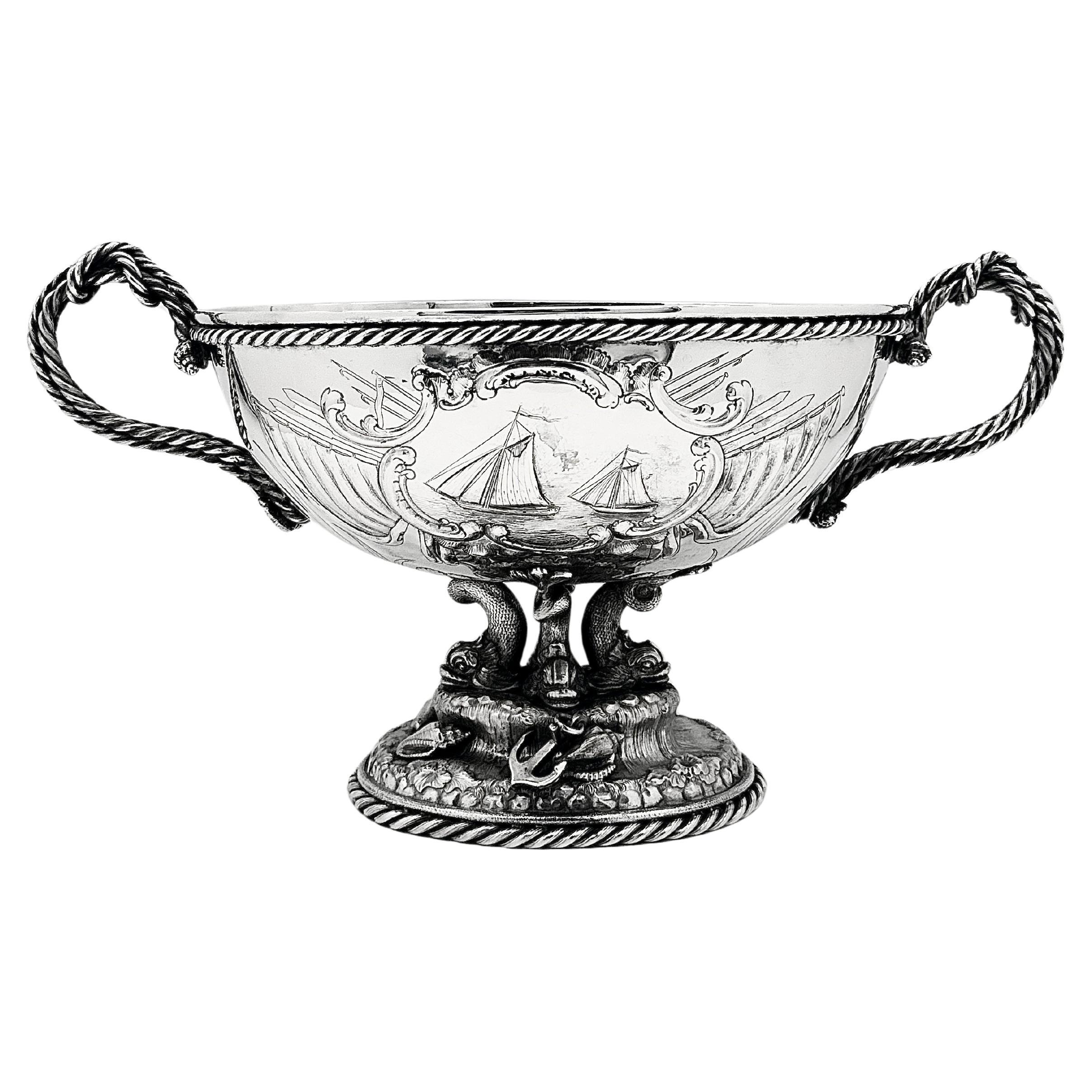 Eine Schale aus amerikanischem Sterlingsilber mit maritimen Motiven, mit einem Rand und einem Fuß mit Seildrehung. Auf dem ovalen Körper sind zwei Rennyachten in einer Kartusche mit Schnörkeln eingeprägt, auf der Rückseite eine leere Kartusche.
Die
