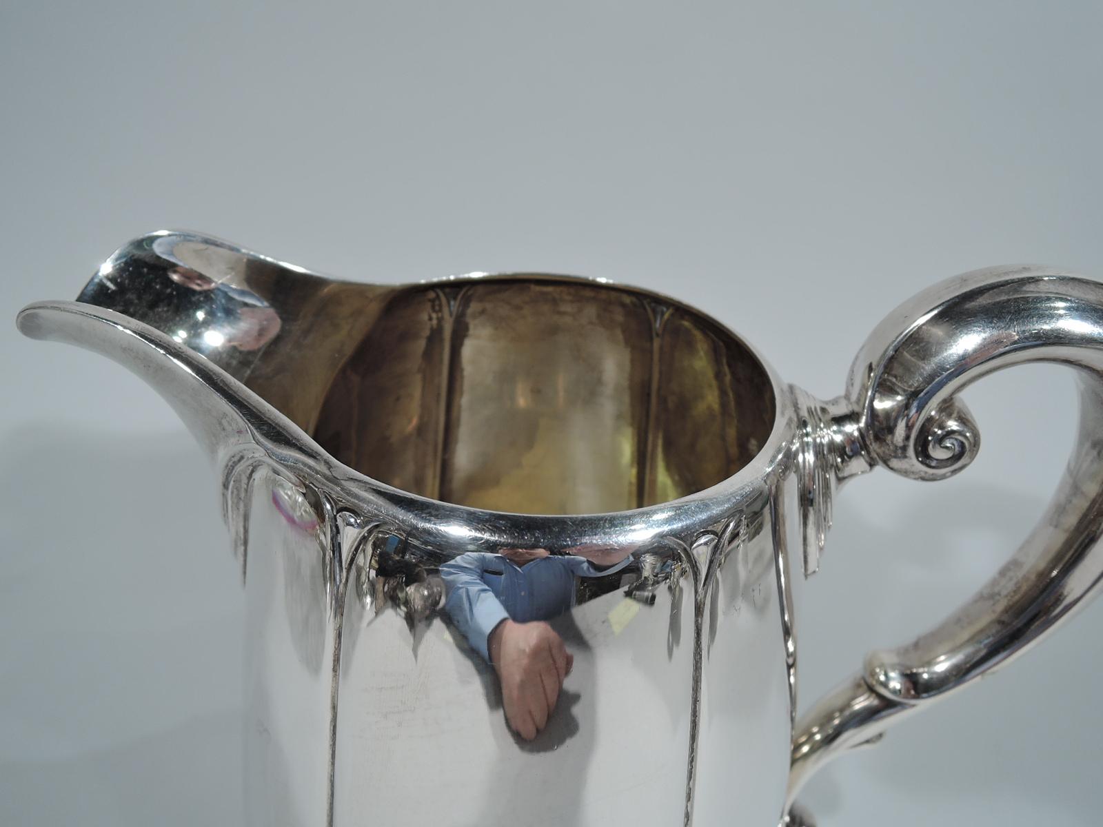 Modern classical sterling silver water pitcher. Made by Dominick & Haff in New York in 1928. Lobed and upward tapering sides, capped foliate scroll handle, wide lip spout with fluid swags, and raised circular foot with crimped trefoils. Sleek but