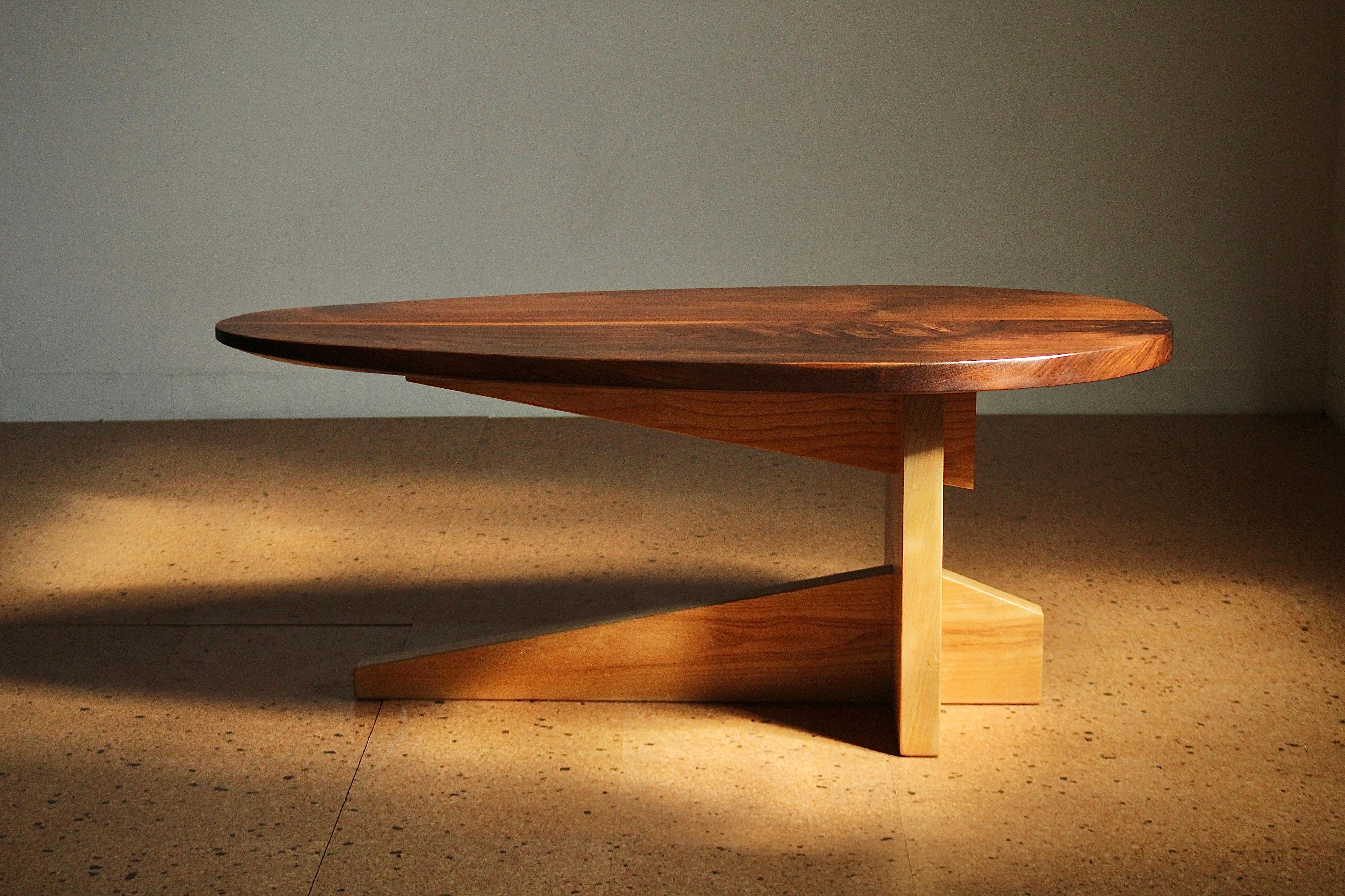 This American studio craft coffee table was likely made in the 1980s-1990s. It features a wonderful egg form top in solid walnut, and a solid oak, cantilevered cruciform base. The thick walnut top gradually tapers, forming a sculpted edge on once