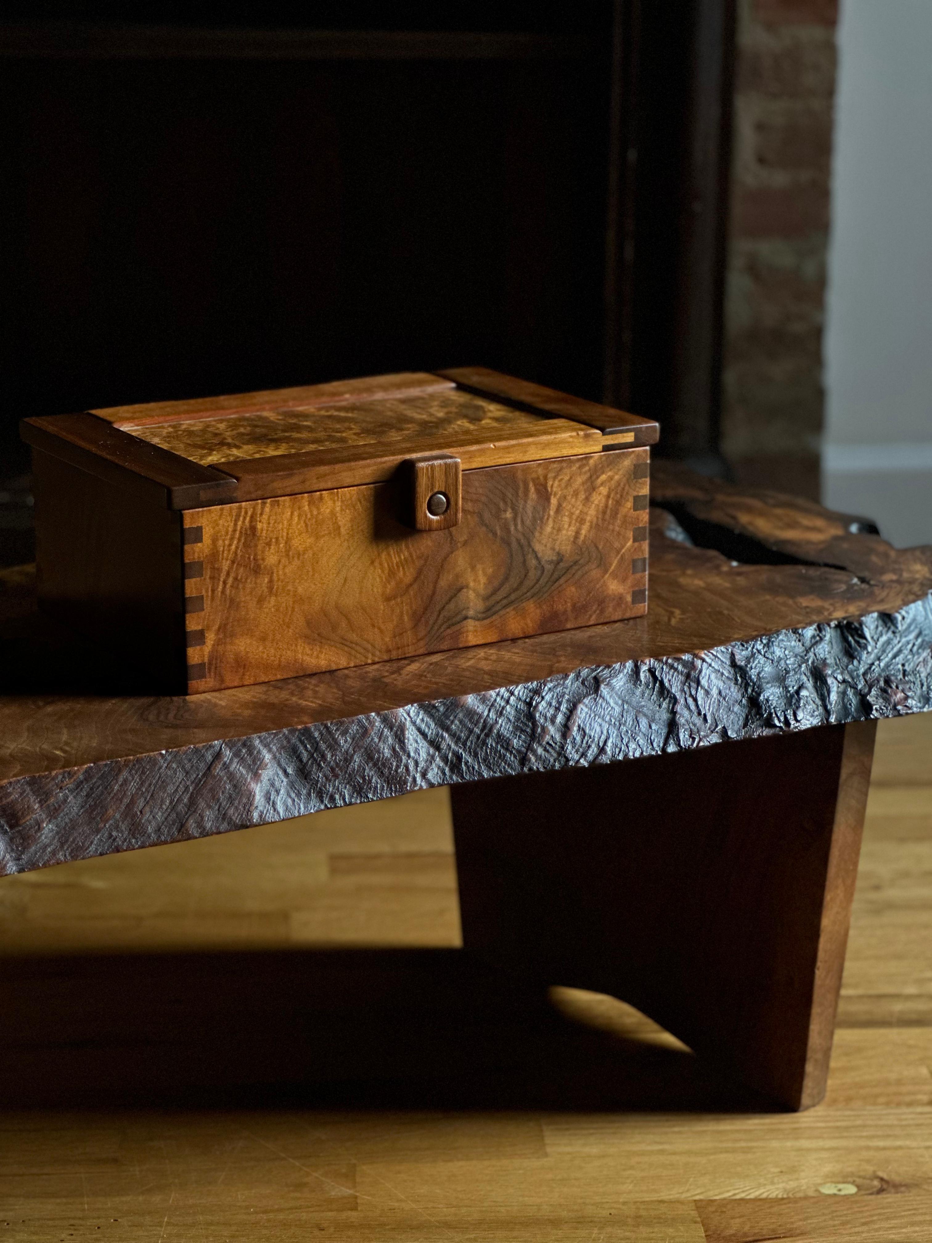 A rectangular box in satin-finished walnut and bird's-eye maple with dovetailing to both body and lid, large wood hinges, and a spring-loaded button closure. Aside from what seem the artist's carved initials 