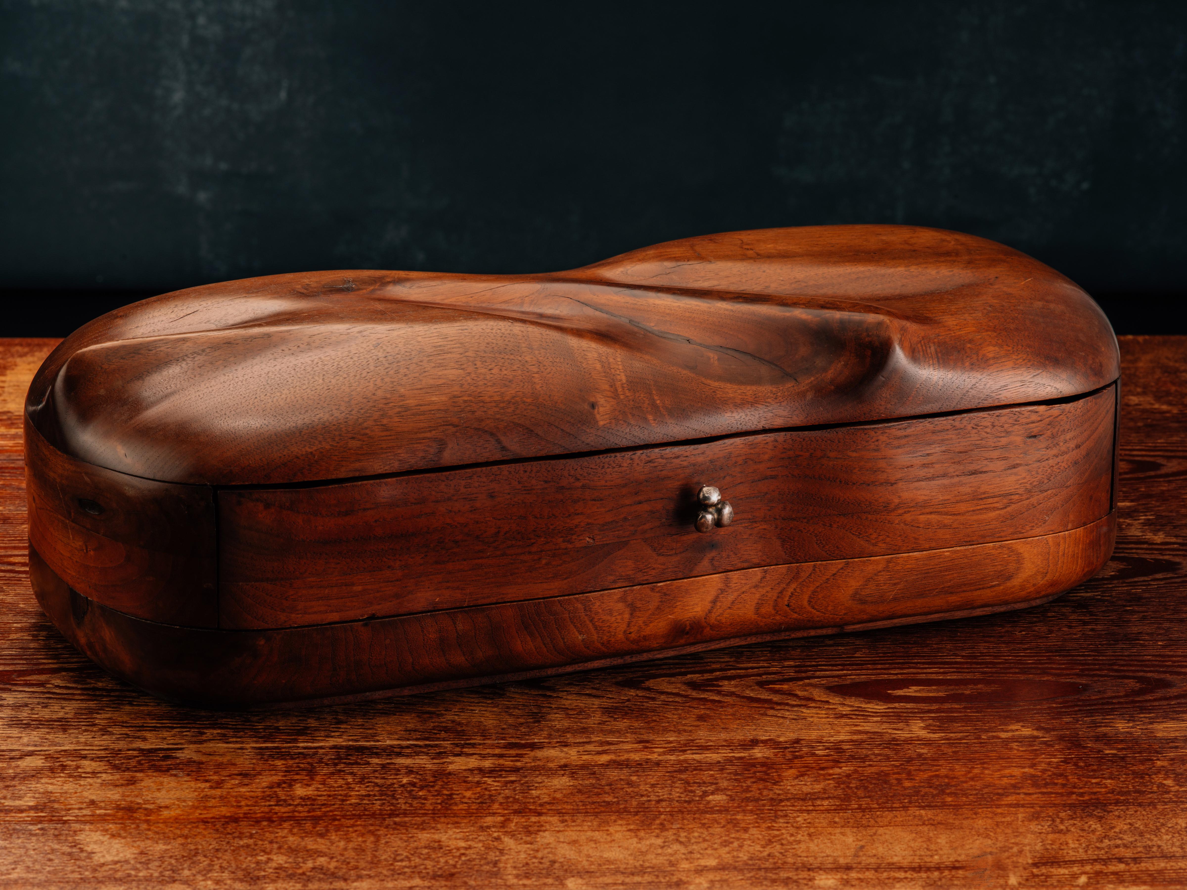 A modernist, walnut jewelry box in a near racetrack form comprising a laminated body, a sensuous, hand-sculpted lid, and a single drawer fitted with a forged metal pull, all raised upon a recessed, shallow foot (3/16