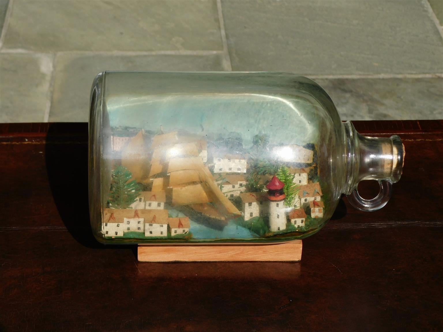 American three masted clipper ship in bottle with cloth rigging in full sail on a painted sea, flanked by a sea side town with evergreen trees, a painted light house on shore, and resting on the original wood stand. Late 19th century.