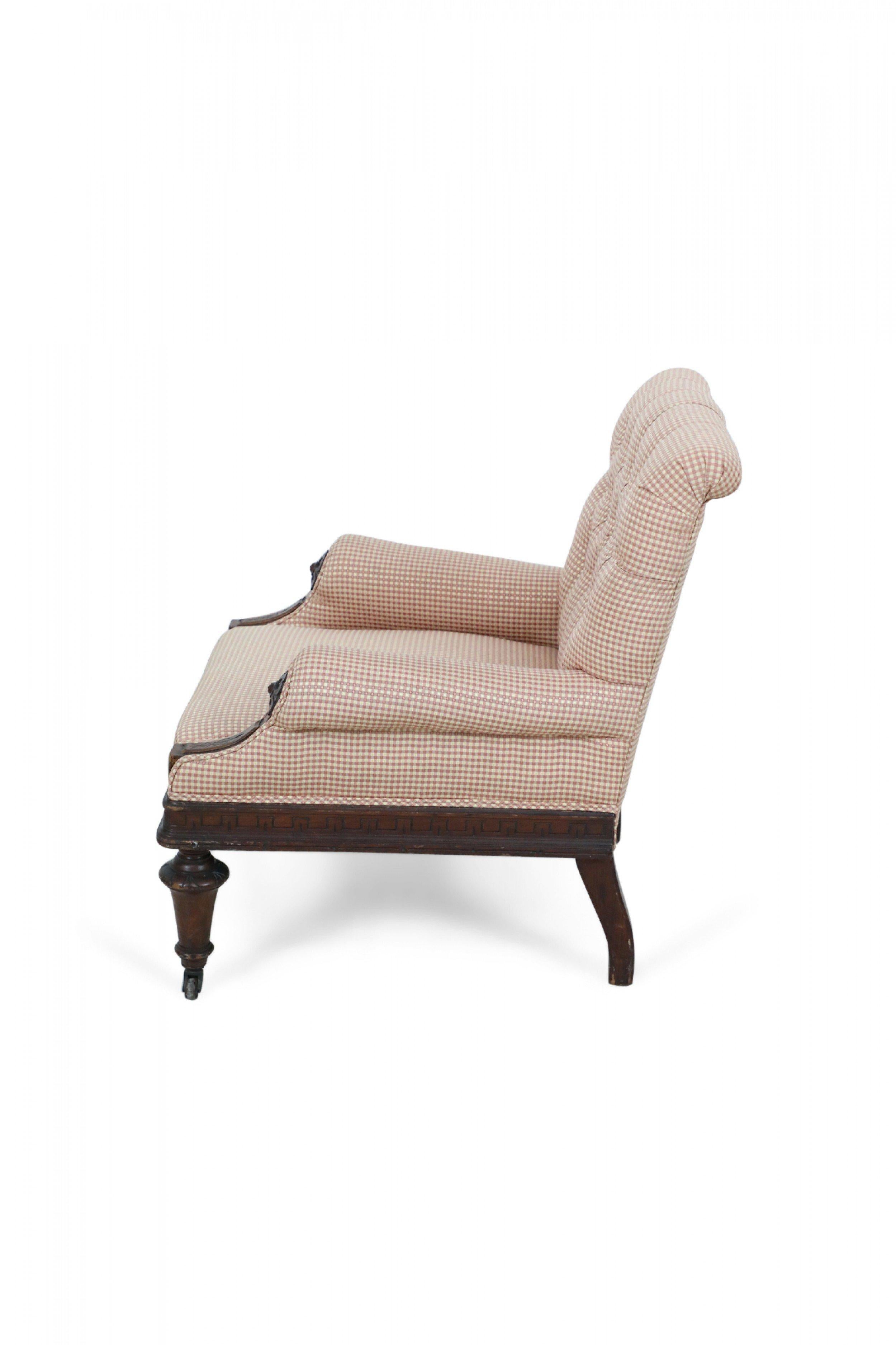 American Victorian Eastlake style armchair with burgundy and beige checkered upholstery with a button tufted roll back and a mahogany frame with carved detail including floral medallions on the arm fronts, resting on brass casters.