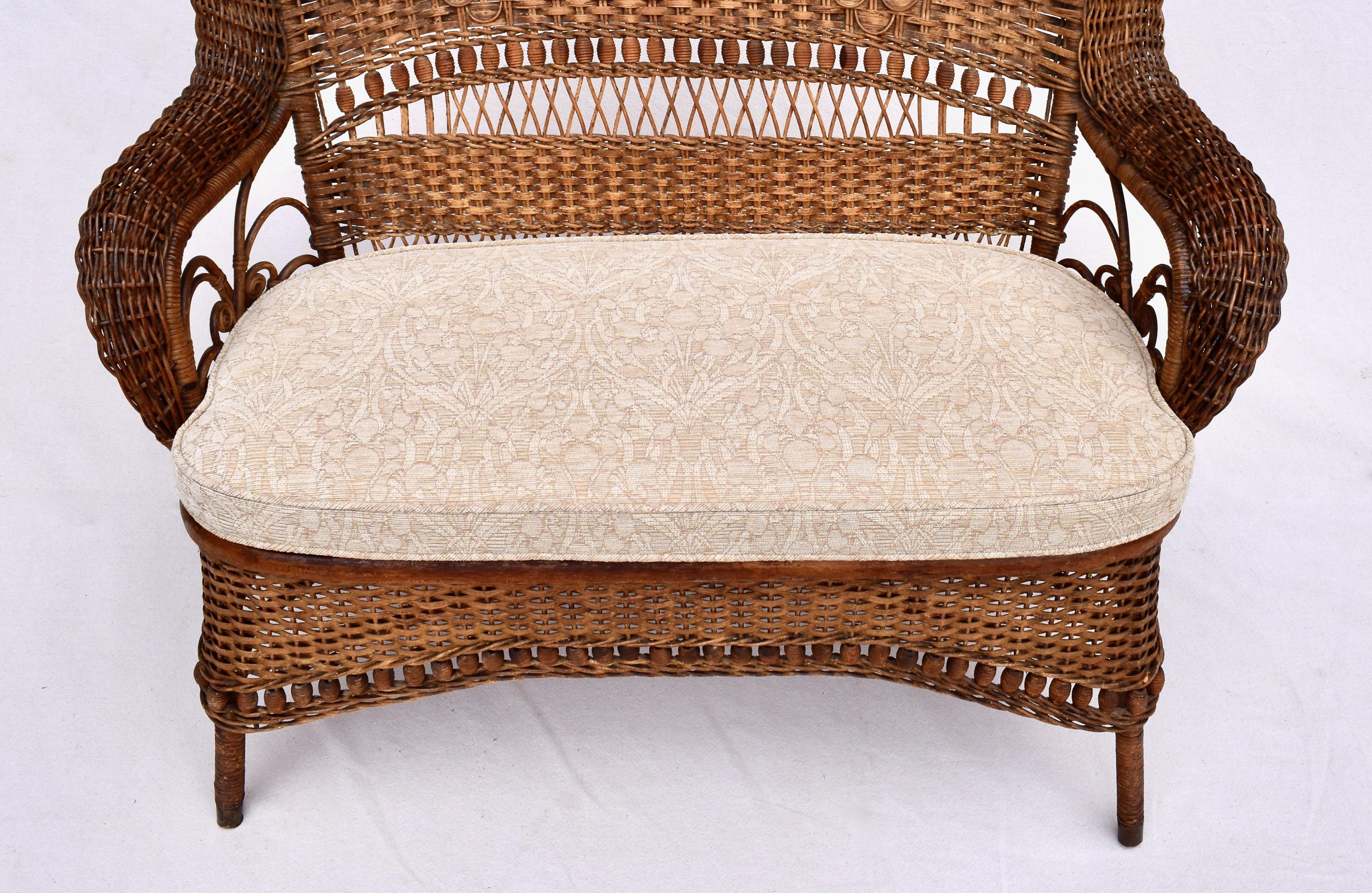 American Victorian natural wicker woven roll design high back loveseat with spindles and bobbins on back and apron. Additionally the settee features new Arts & Crafts upholstered custom cushion & caned seat. Brass capped feet & original tag remnants