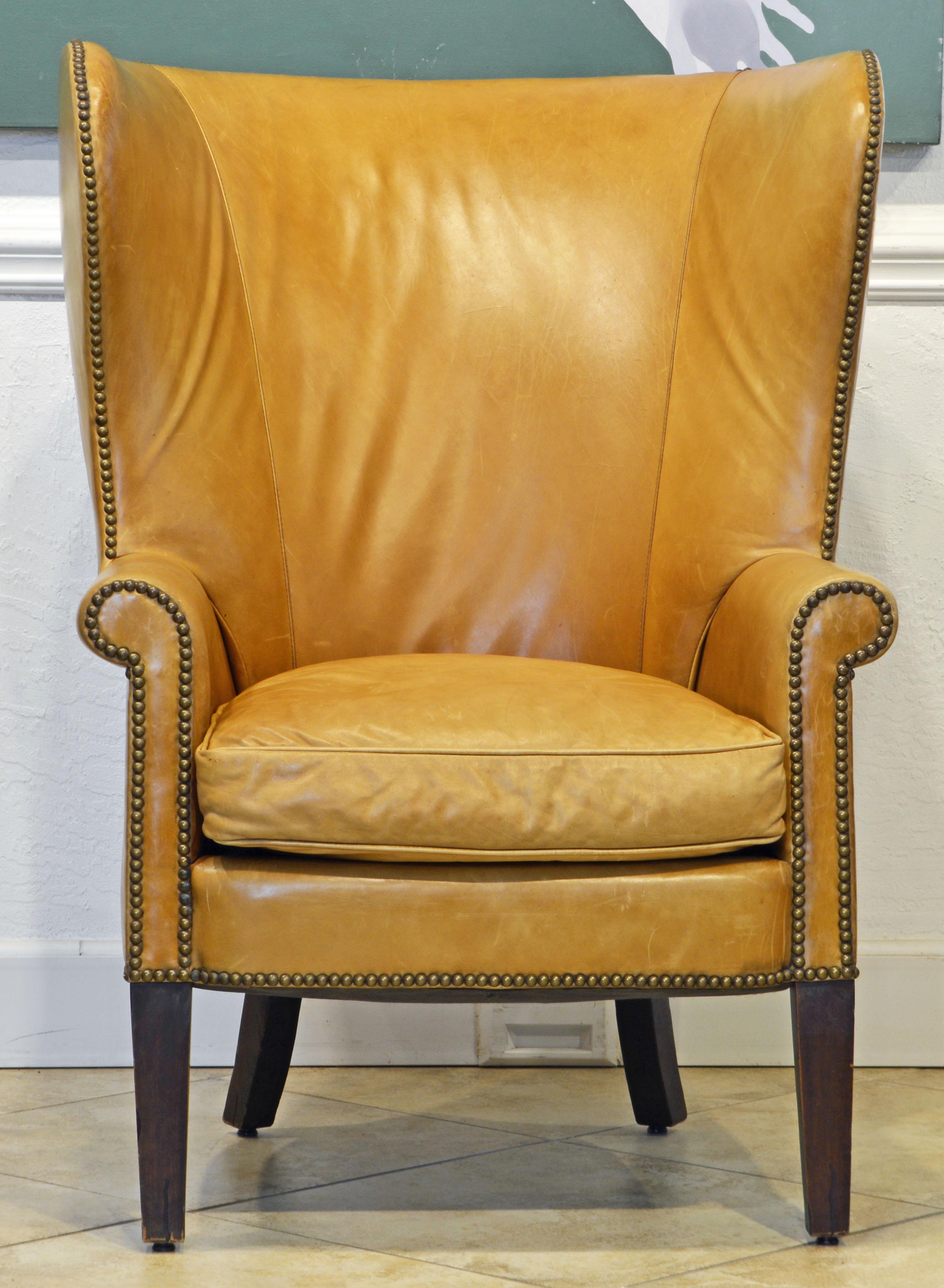 This sculptural wing back chair feature tanned leather cover trimmed with brass nail heads. It is a comfortable and inviting chair that will add an English country house flair to an interior.