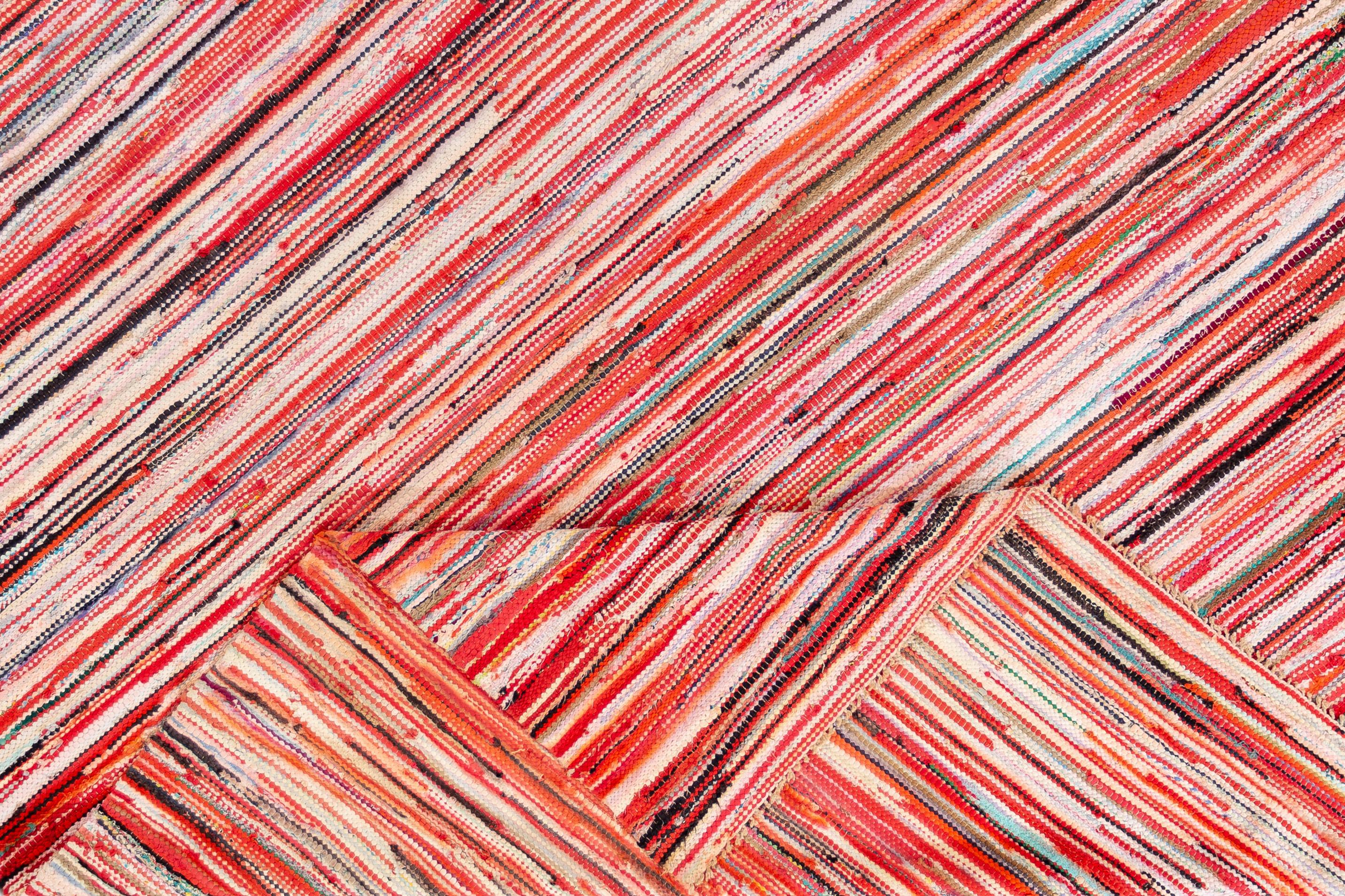 A handwoven American Rag rug. This rug has a bright, multicolored design in thin, horizontal stripes (primarily red and orange). This rug measures 9'4