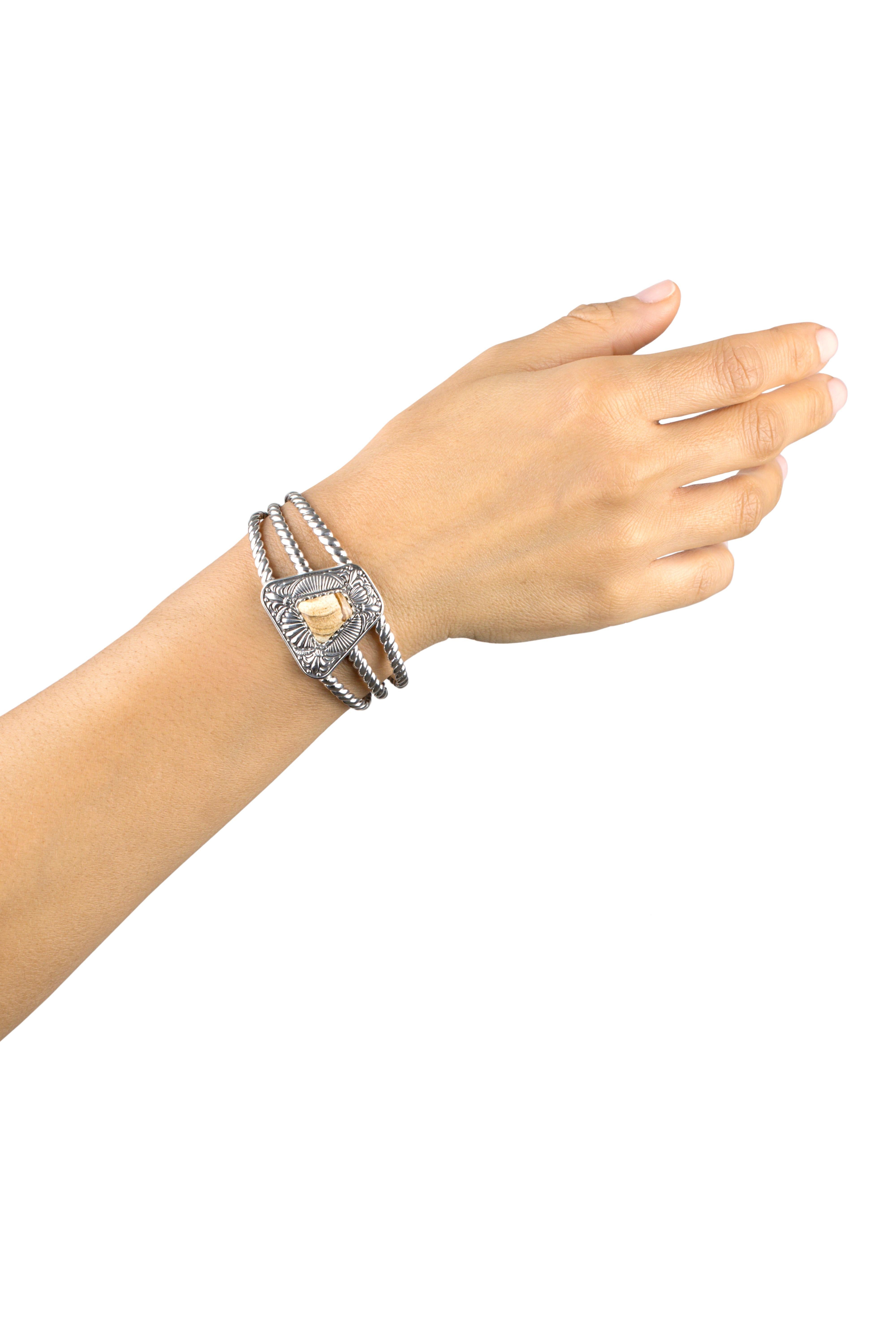 - Pietra preziosa di diaspro tagliata a forma di trapezio e incastonata in un castone dentellato su un ampio bordo in argento sterling ossidato con accenti floreali e a ventaglio.
- Polsino aperto in argento sterling, a tre file, con dettagli in