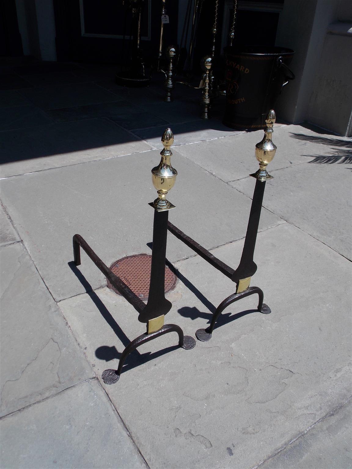 American wrought iron and cast brass flanking urn finial knife blade andirons with scrolled legs and penny feet. The bulbous iron plinth is more vigorous and bolder than most with an additional decorative brass shied to cover blacksmiths joint
