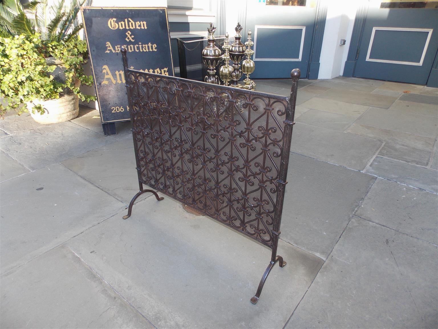 American wrought iron free standing fire screen with flanking ball finials, decorative scrolled centered panel with interior lined mesh, tapered supporting columns, and terminating on scrolled legs with stylized penny feet, Early 19th Century.