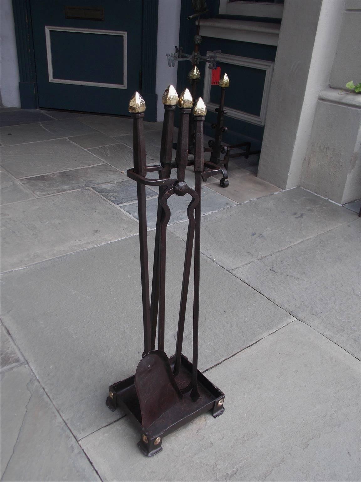 American wrought iron and brass fire tools on stand with faceted arrow finials, tapered squared shafts, artistic scroll work, and terminating on a decorative rectangular base with corner brass medallions and stylized hammered feet. Set consist of