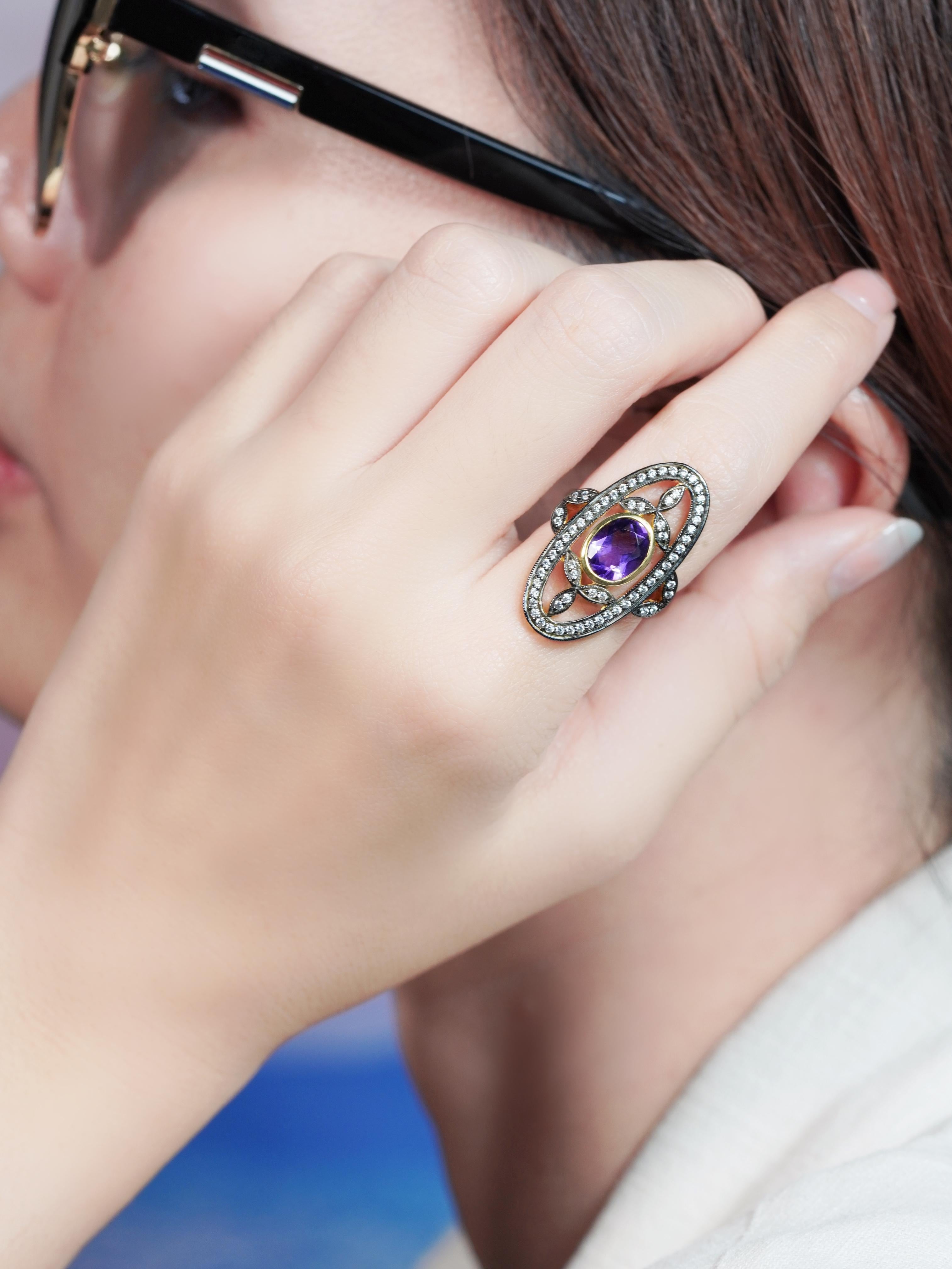 El anillo floral de estilo antiguo muestra una impresionante amatista ovalada en su centro, que brilla con ricos matices púrpura. Flanqueado por delicados detalles de diamantes inspirados en los pétalos en flor, el diseño capta maravillosamente la