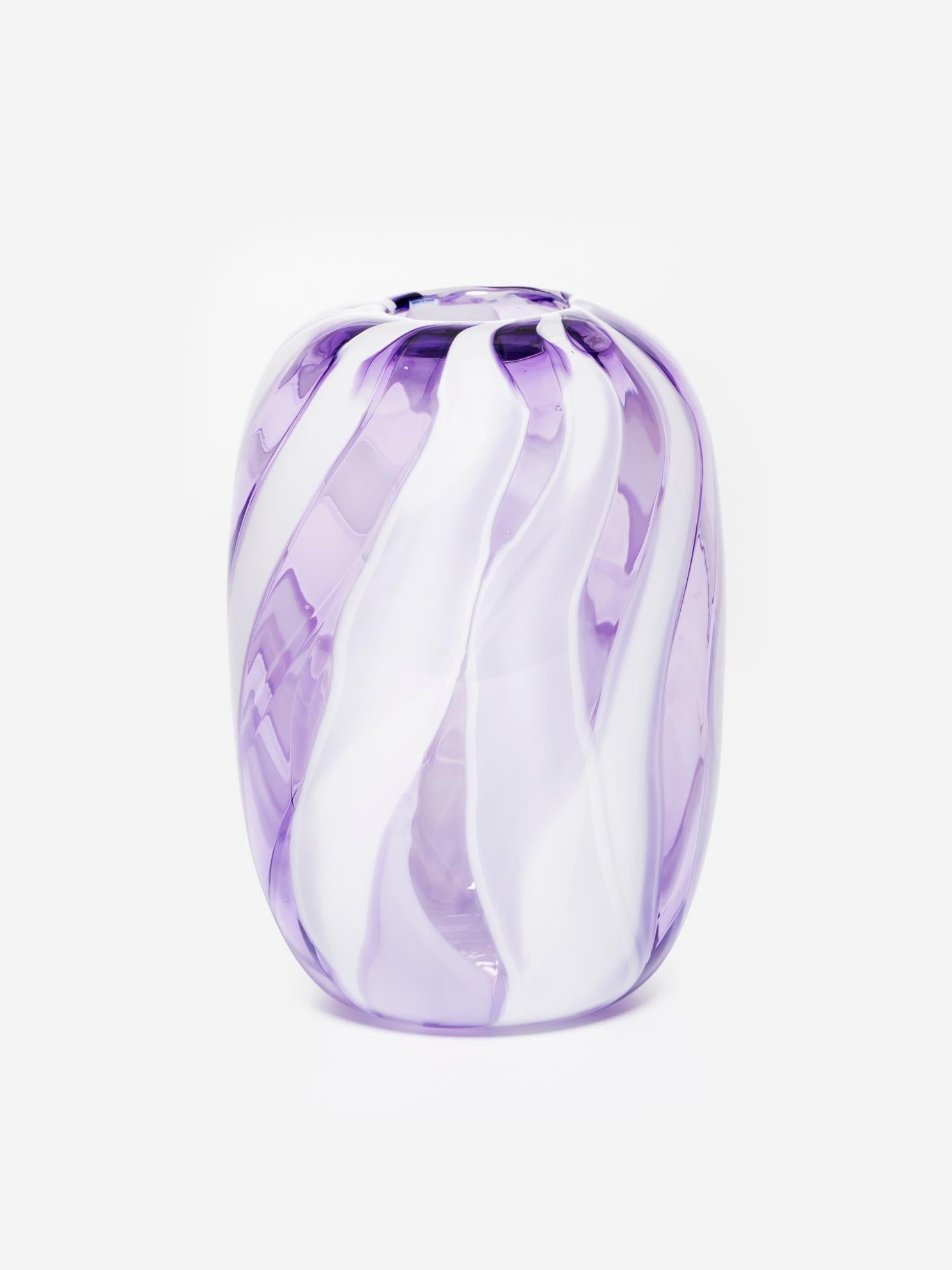 Per questo bellissimo vaso di vetro a forma di bocciolo, un team di soffiatori di vetro ha prima estratto canne di vetro bianco e ametista. Una volta fredda, la canna viene tagliata in bastoncini e disposta a strisce, per poi essere nuovamente