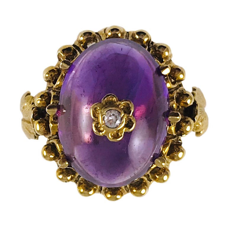 14 Karat Yellow Gold Amethyst Diamond Ring. The Amethyst Cabochon measures 16 x 13mm and is six-prong set. Six small gold petals form a flower at the center of the cabochon with the center of the flower featuring a bezel-set round diamond. The ring