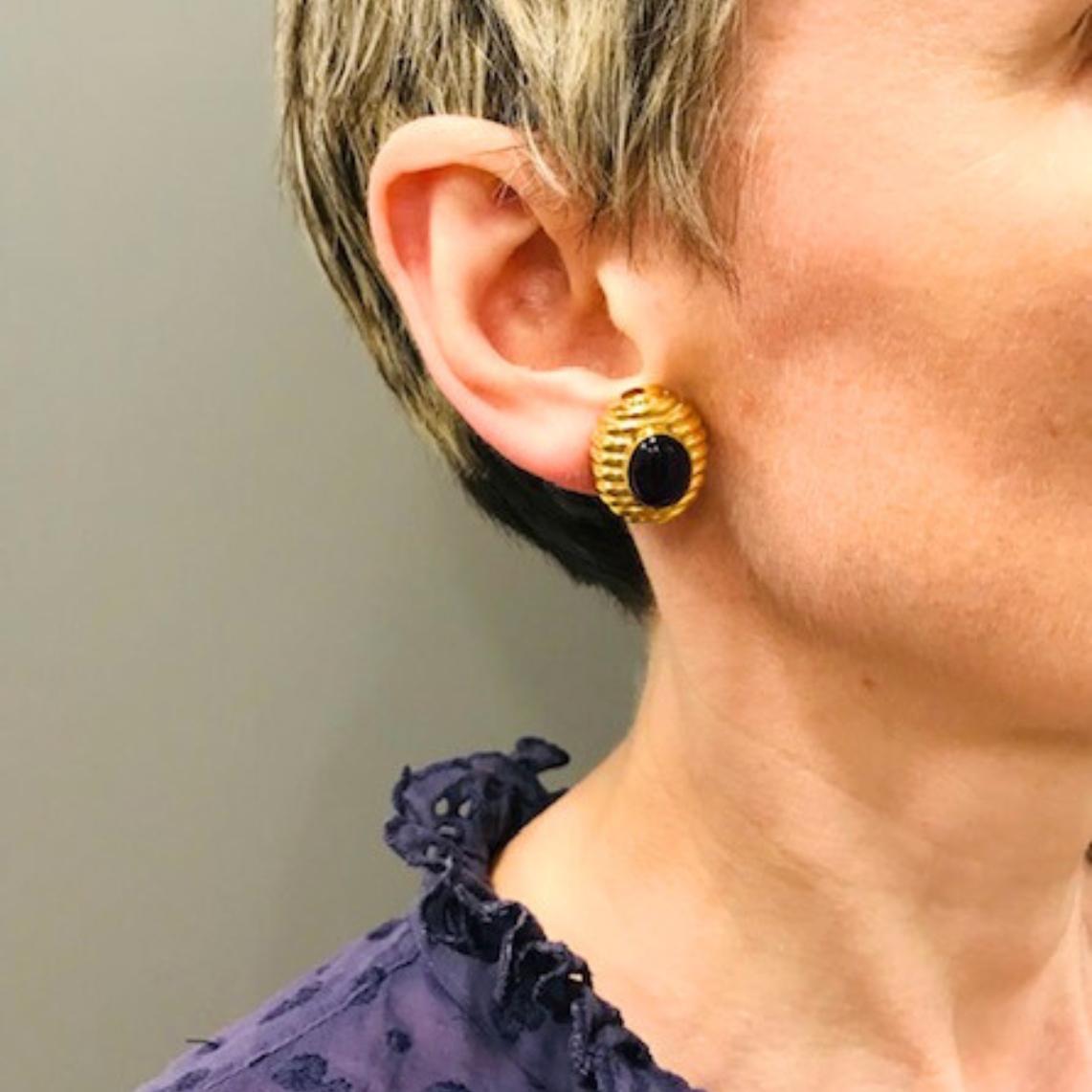 Un paio di orecchini a clip vintage di Tambetti, realizzati in oro giallo 18 carati e dotati di ametiste ovali tagliate a cabochon. Ogni orecchino ha una montatura d'oro a gradini. La sua struttura è coerente e simmetrica, con le scanalature che