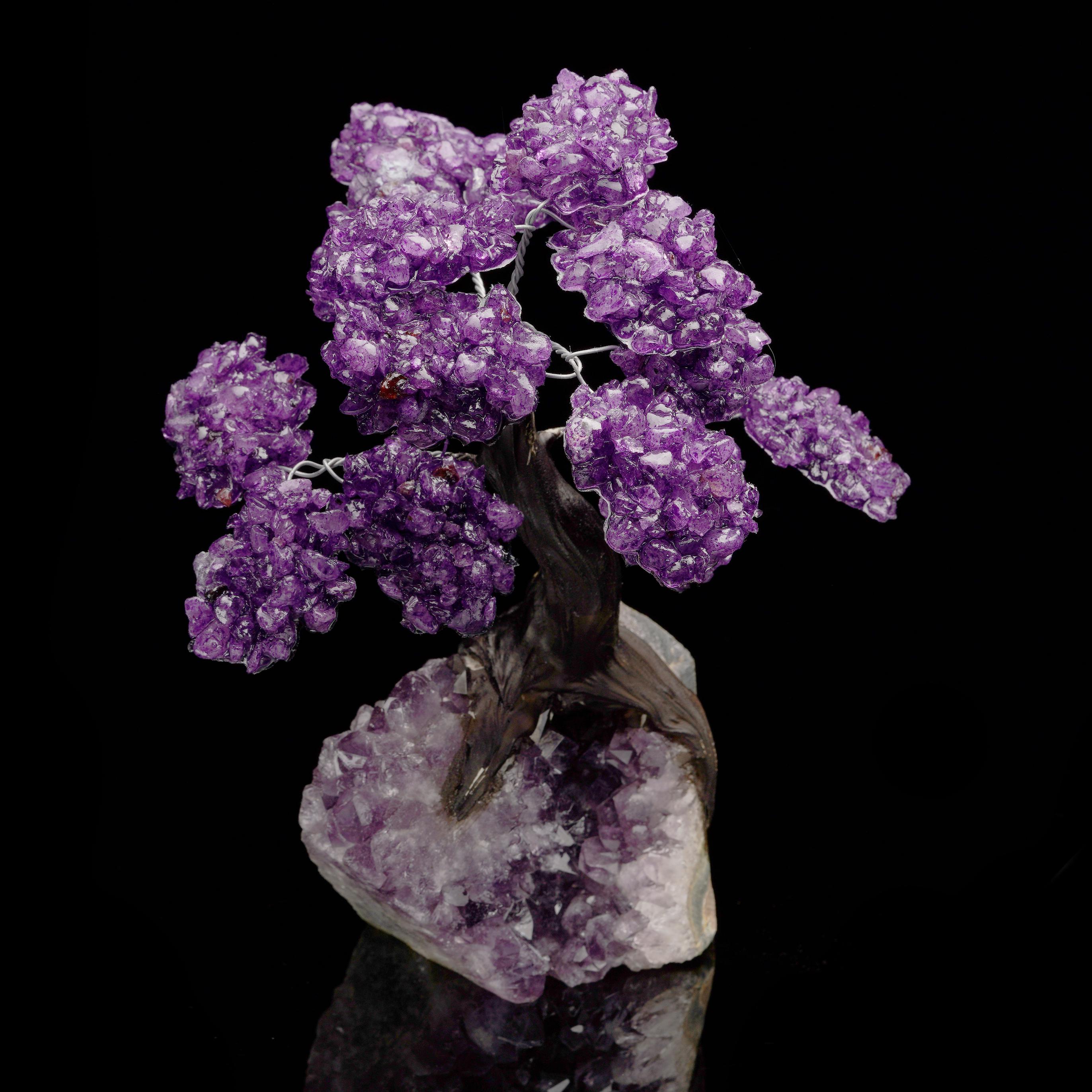 Dieser handgefertigte Edelsteinbaum zeigt leuchtend violette Amethystkristallblüten, die an einem anmutig gewundenen dunklen Stamm blühen, der elegant in einem natürlichen Amethystcluster verwurzelt ist. Die auffällige Kombination aus dem