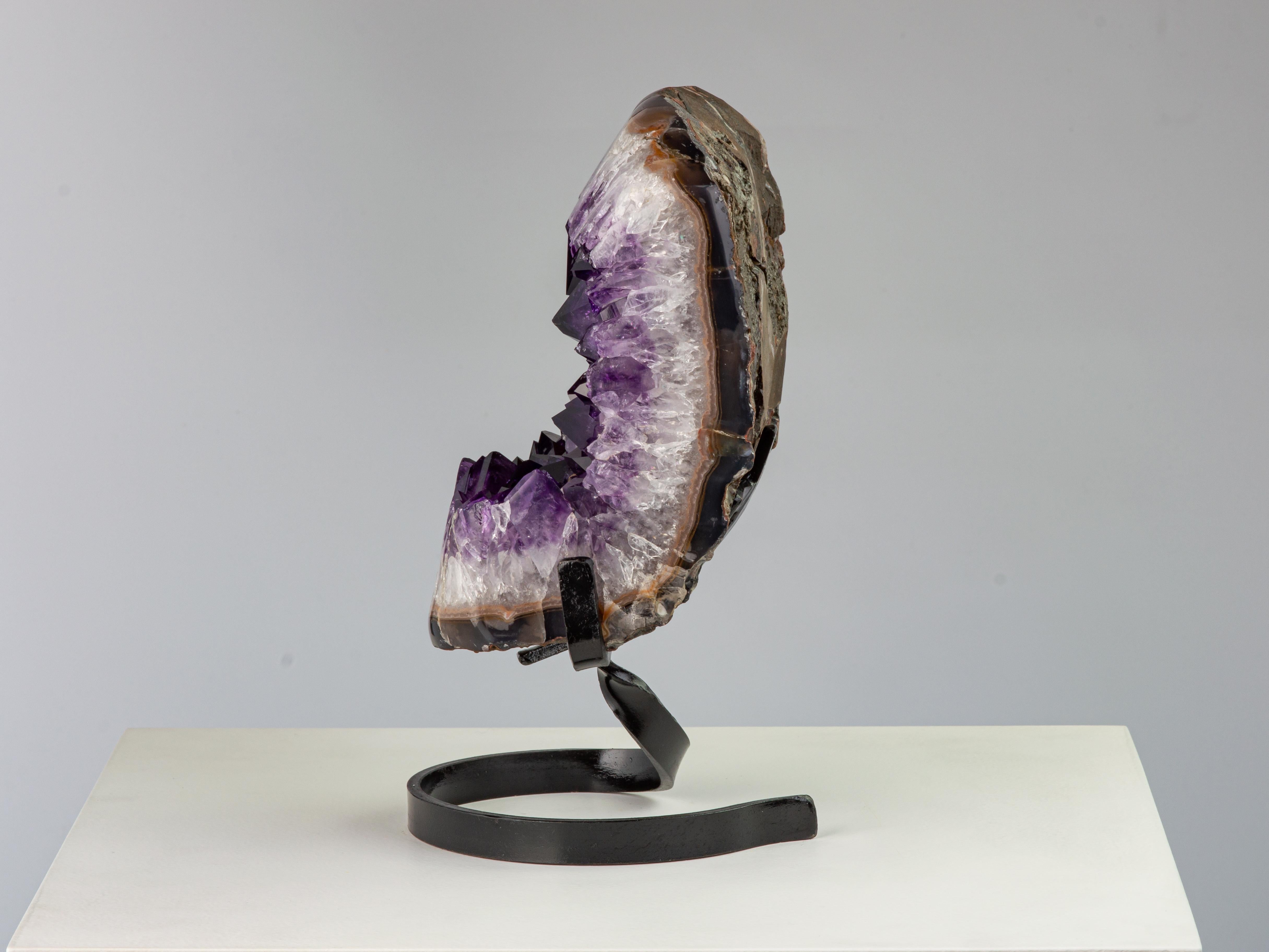 A stunning deep purple amethyst cluster with unusually high peaked crystals on metal stand. The polished frame shows mineral layers developing from the rough basalt, to the grey/blue agate, jasper, a thick layer of white quartz and finally to the