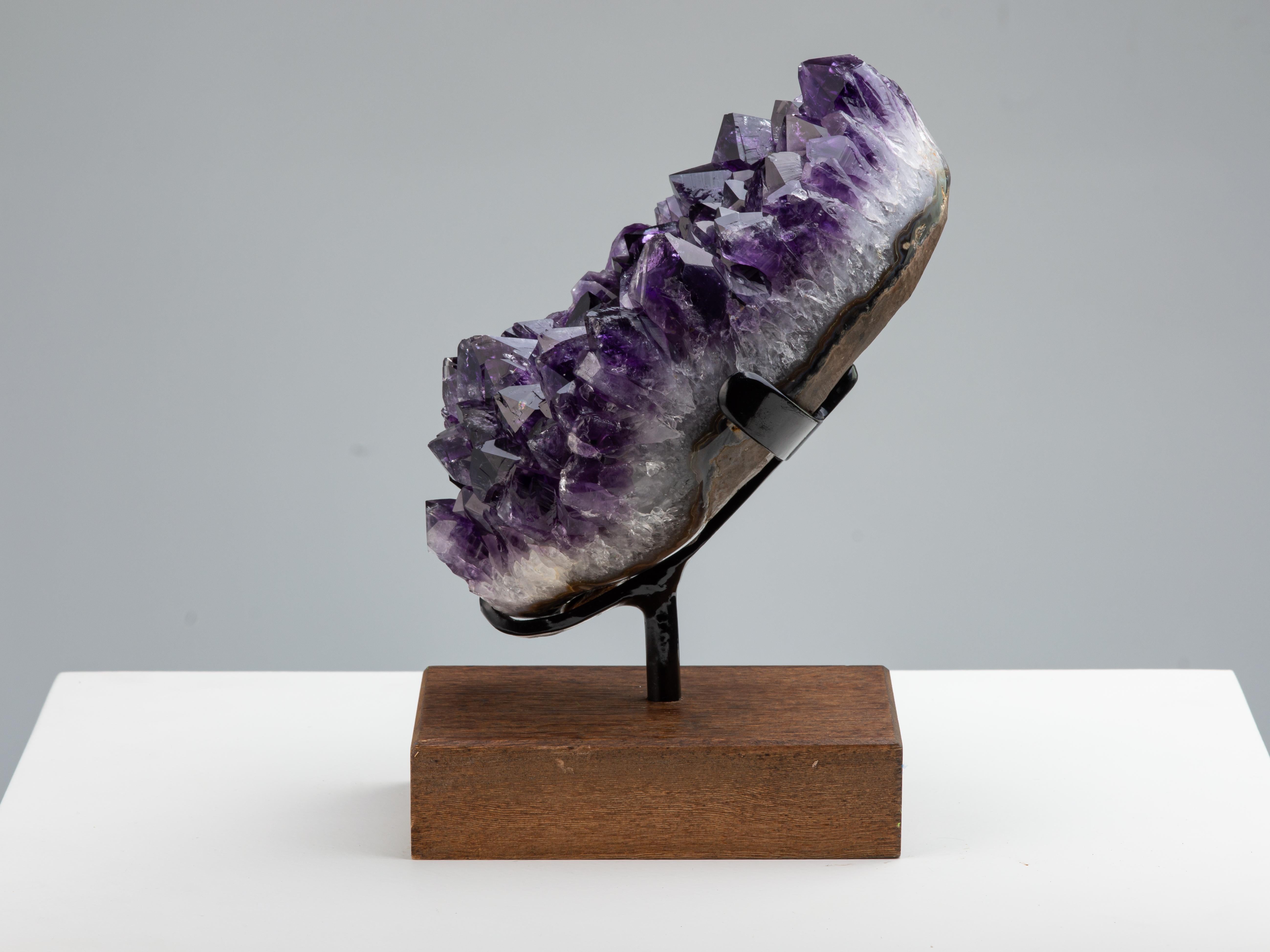 Une magnifique grappe d'améthyste d'un violet profond avec des cristaux à hauts sommets. Le cadre poli montre des couches minérales se développant à partir du basalte brut, de l'agate grise/bleue, de l'épaisse couche de quartz blanc et enfin du