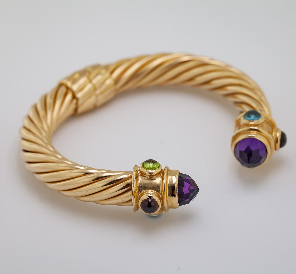 The 14k yellow gold cable twist hinged mounting, 
9.5 mm in width, both terminals feature 
(1) bezel set faceted top amethyst cabochon, 
8.8 X 6 mm, accented by (2) 4.5 mm, 
bezel set round blue topaz, garnet and peridot cabochons, 
internal