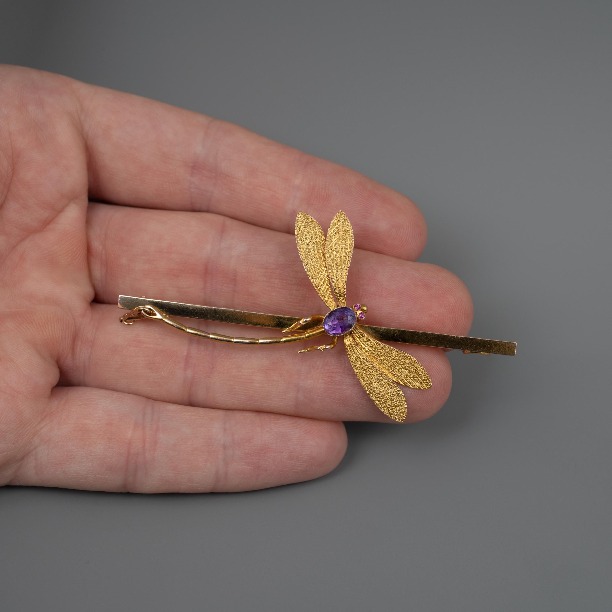 This outstanding Dragonfly brooch pin, set with amethyst and rubies is beautifully crafted in 9 karat yellow gold. The dragonfly is nicely positioned onto a squared bar and features textured finish to the double wings. The gold pin slots neatly into