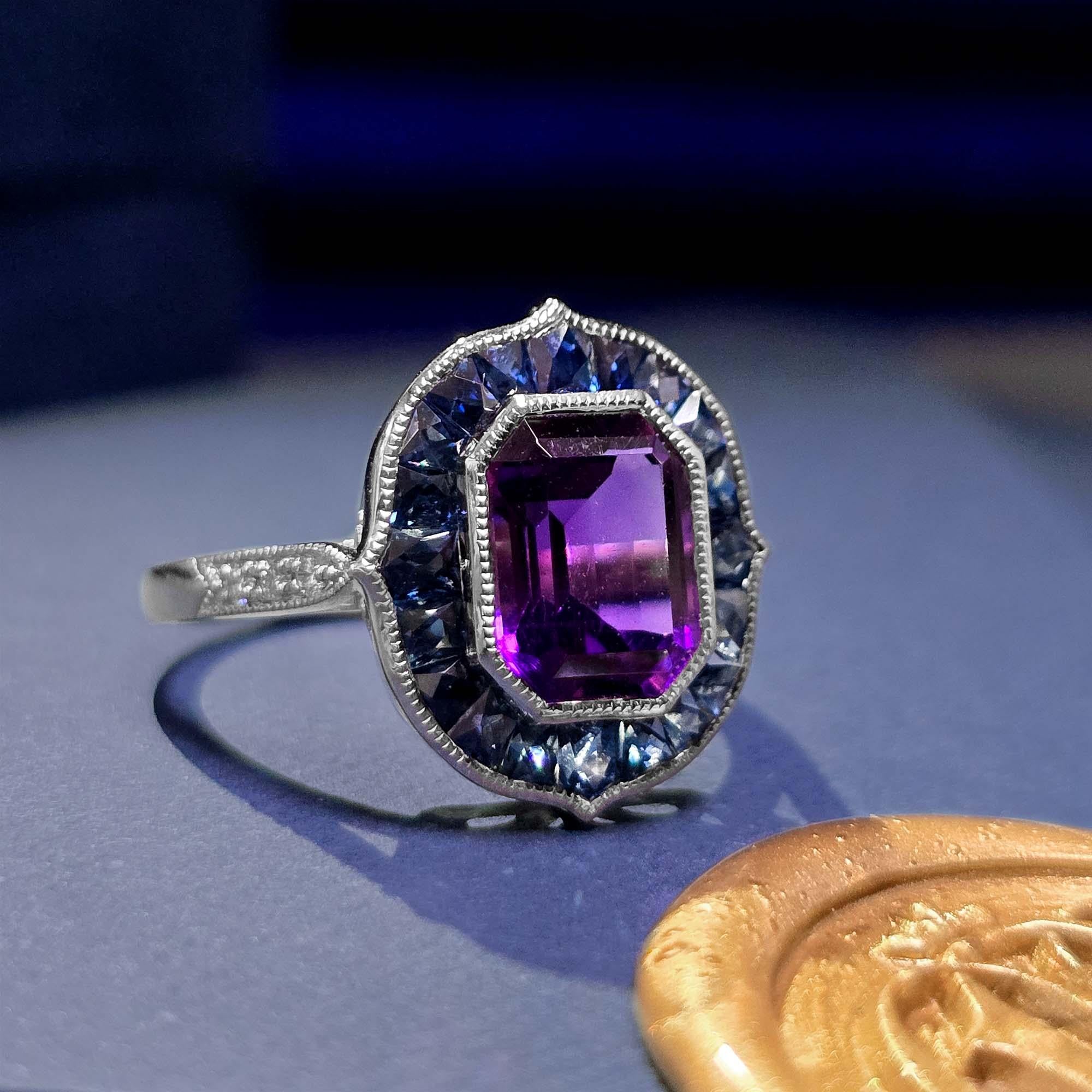 A striking emerald cut amethyst at the center, surrounded by a rich sapphires and sparkling diamonds on the shoulders. The geometric arrangement of sapphires and diamonds enhances the deep purple hue of the amethyst, creating a bold and